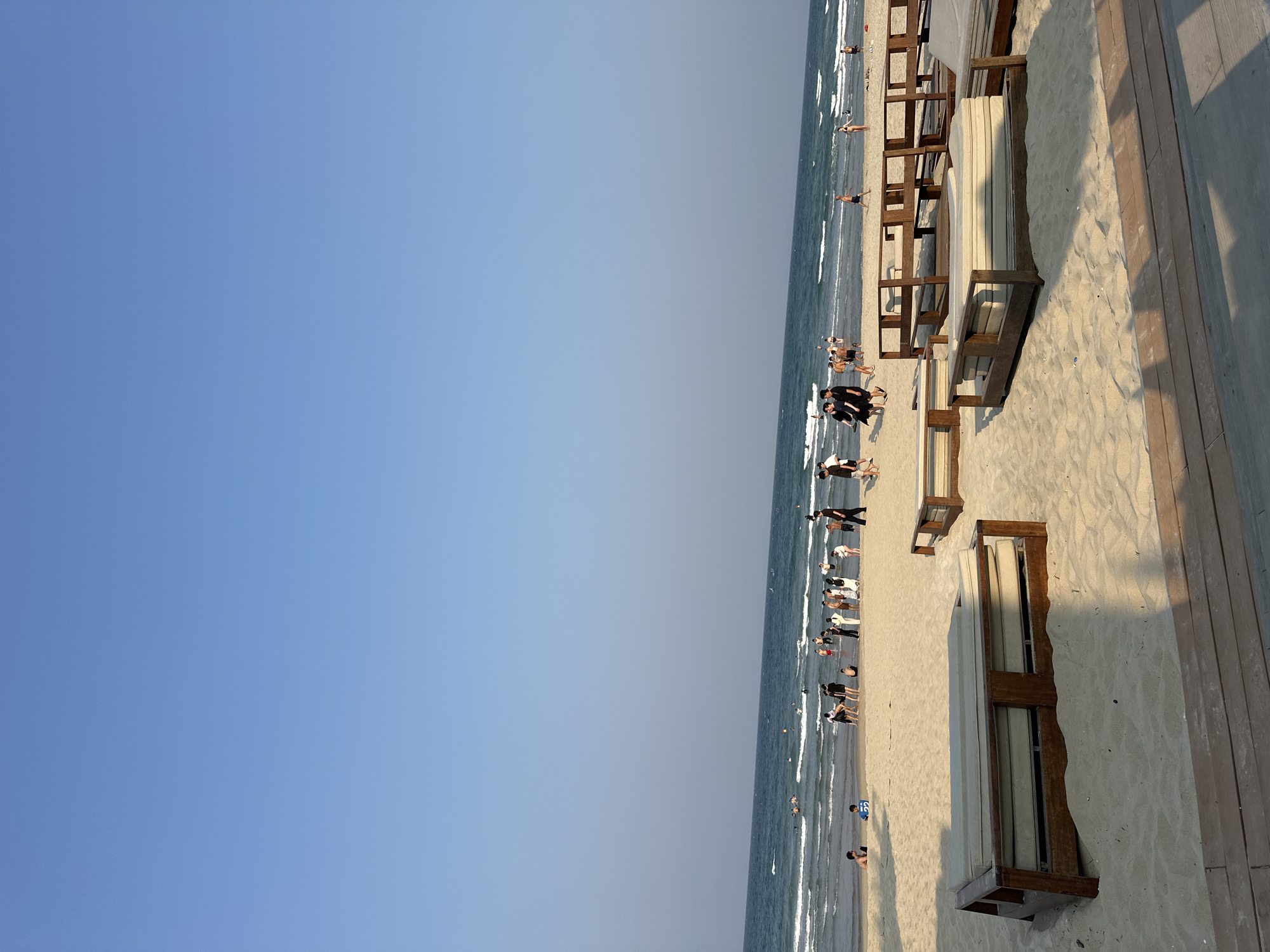 The image depicts a beach scene, possibly in Đà Nẵng, Vietnam. The sky is a clear blue, taking up a significant portion of the frame. The ocean is visible, with small waves breaking on the shore. There are people along the beach, some walking along the shoreline and others in the water. Beach chairs, possibly for rent, are arranged on the sand. A wooden walkway is present along one edge of the beach, potentially separating it from a building or a resort.