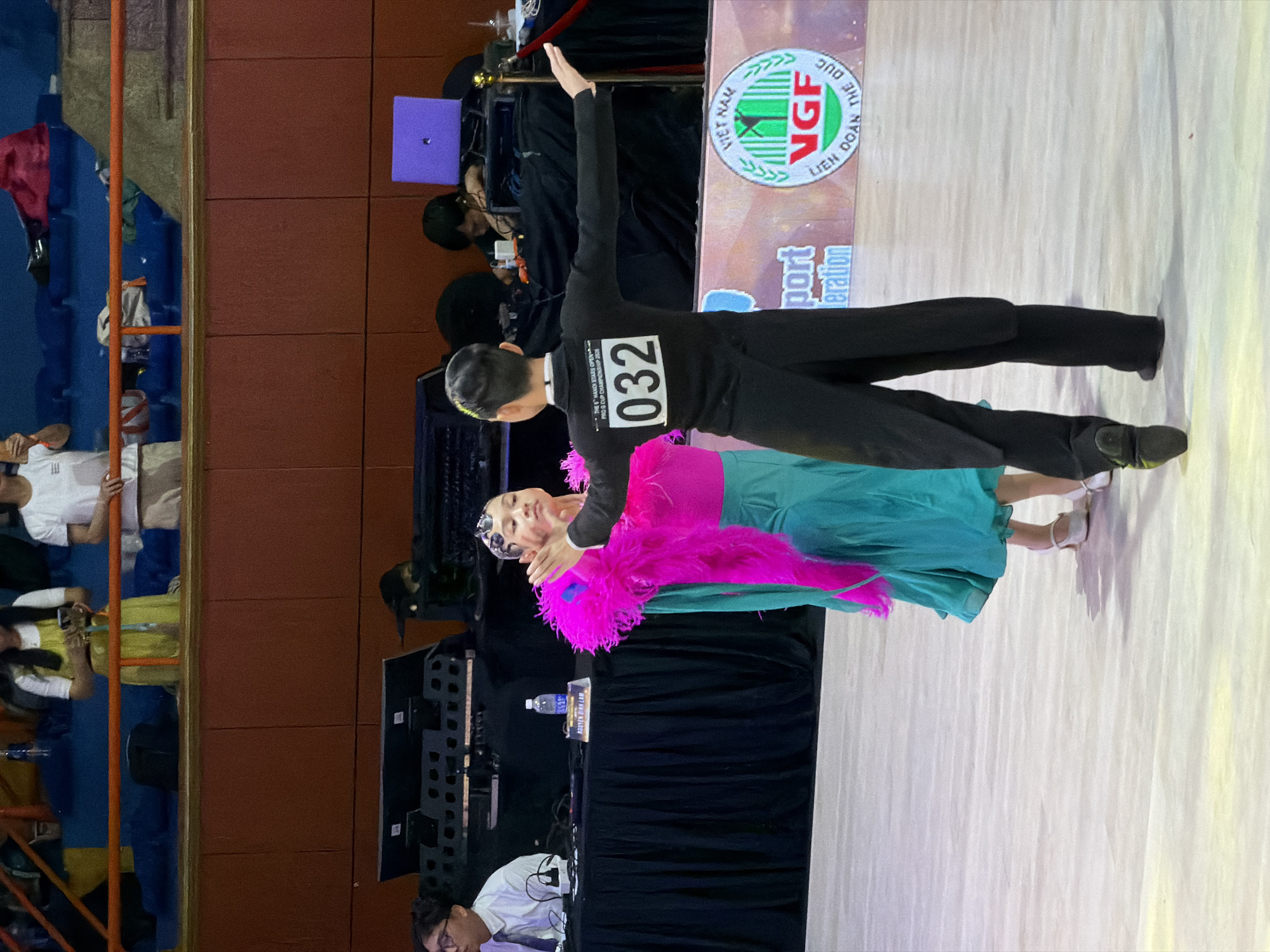 The image depicts a ballroom dancing competition, likely held indoors in a venue with tiered seating. Two young dancers, a boy and a girl, are the primary subjects, positioned on a polished floor. The boy, wearing a black tailcoat and pants with a bib bearing the number "032," is facing away from the camera, arms extended in a dance pose. The girl, in a vibrant pink and teal costume, is facing towards the boy, her body turned slightly. The background reveals a stage area with equipment, including what appears to be musical instruments and a laptop. Spectators are visible in the upper tiers of seating. A banner with Vietnamese text and the "VGF" logo, along with "VIET NAM" and "LIEN DOAN THE DUC," suggests the event is taking place in Vietnam, potentially Hà Nội given the presence of Vietnamese branding. The lighting appears to be artificial, illuminating the dance floor and creating shadows.