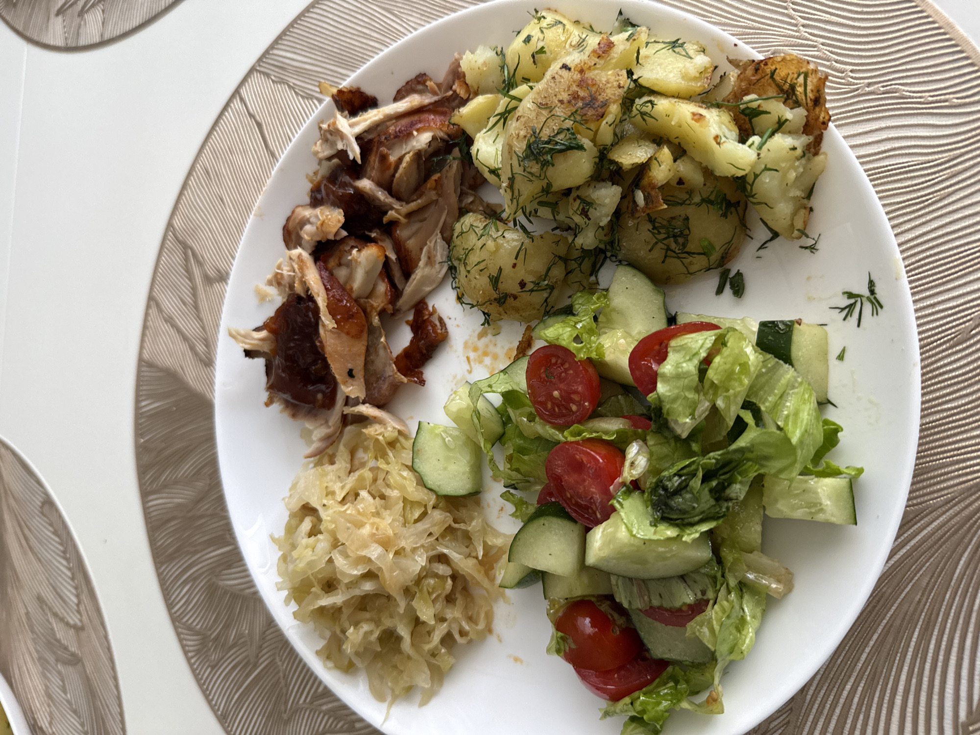 A white plate of food sits on a beige placemat with a woven pattern. The plate contains shredded cabbage, pulled meat with a dark sauce, a salad of lettuce, cherry tomatoes, and cucumber, and roasted potatoes with dill. The food appears to be a meal. The background is out of focus, but shows a surface that could be a table. There are no people or animals visible. The image is taken from a high angle, looking down at the plate of food. The lighting is bright and natural, suggesting it is daytime.