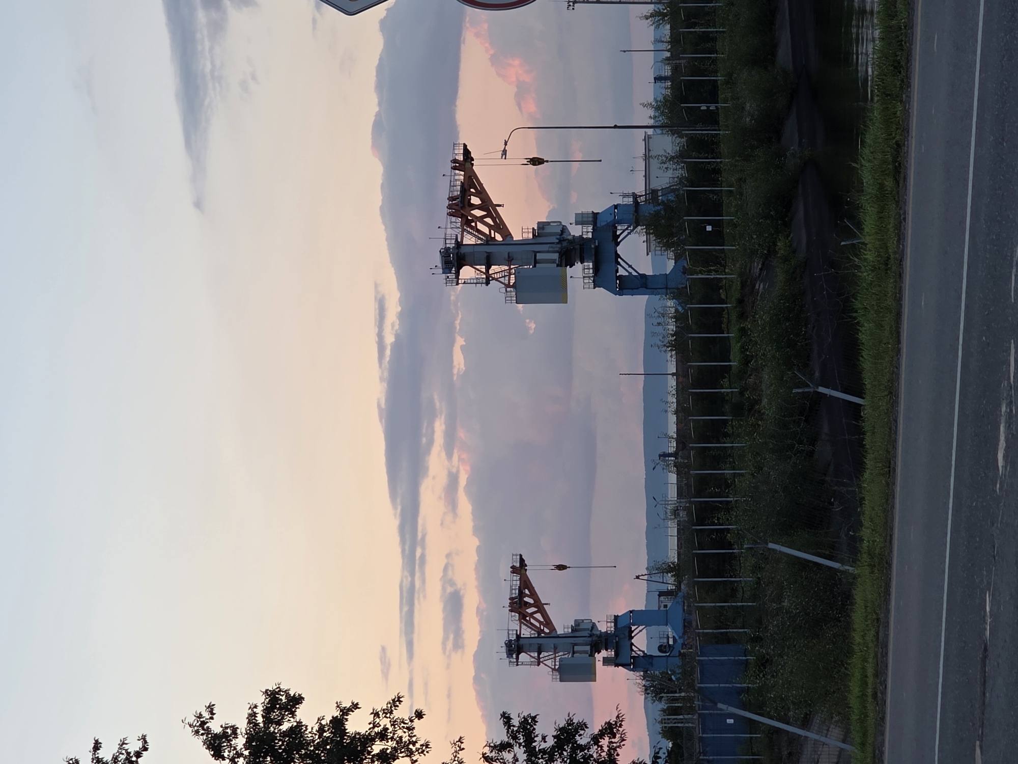 The image depicts a waterfront industrial area at dusk, likely in the city of Chaikovsky, Russia. Two large industrial cranes, painted blue with orange accents, stand prominently against a sky filled with soft pink and purple clouds. The cranes are positioned on a waterfront, with a body of water visible in the background, beyond which lies a distant shoreline with hills or a treeline. In the foreground, a paved road runs horizontally across the bottom of the frame, separated from the industrial site by a grassy verge and a chain-link fence. The industrial area itself is characterized by various structures, including buildings, a fence line, and several lampposts. Vegetation, including trees and bushes, is present throughout the scene, some of which partially obscure the industrial elements. There are no people visible in the image. The overall atmosphere is calm and serene, with the soft lighting of dusk creating a muted color palette.