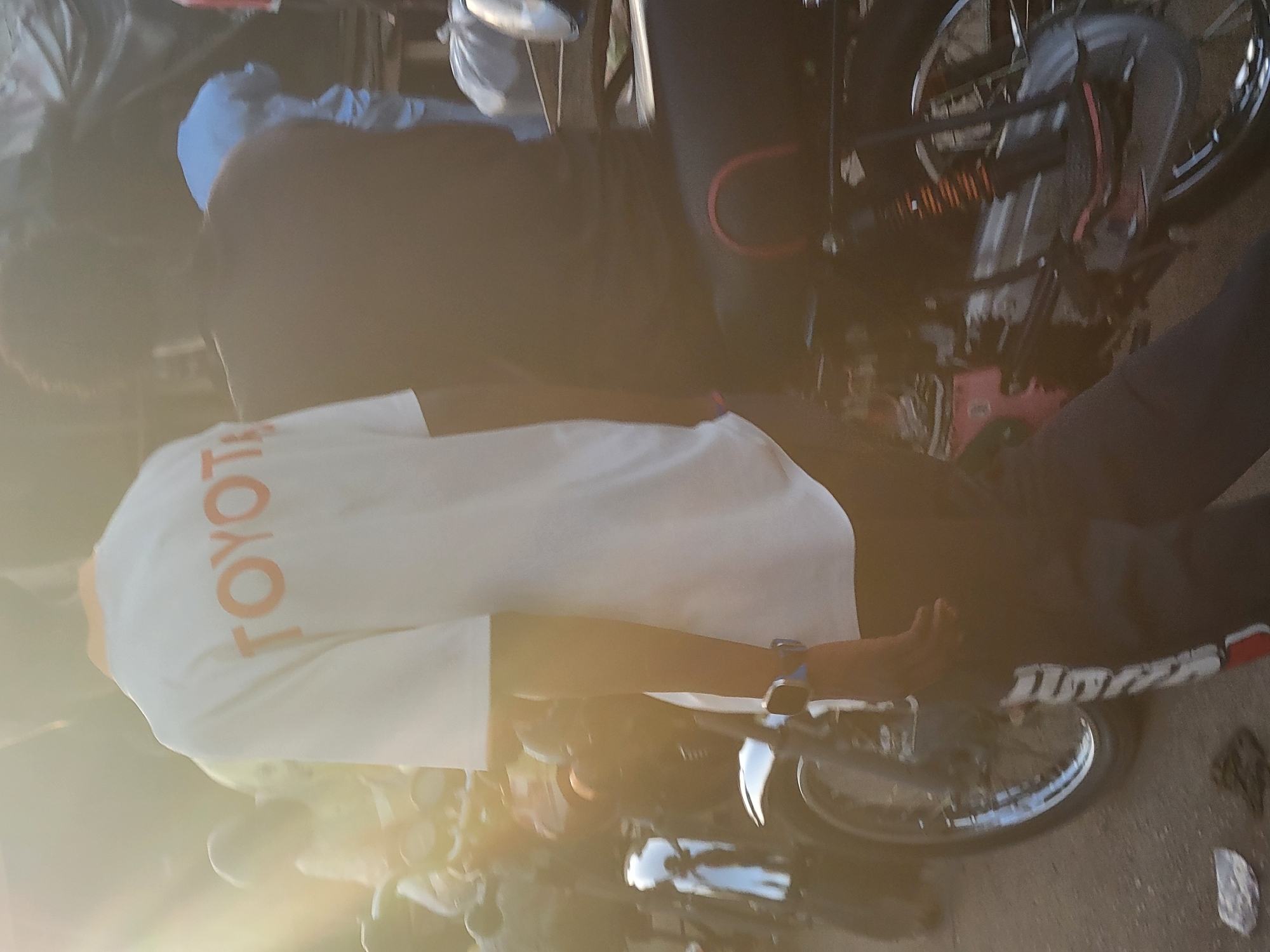 The image depicts a person wearing a white shirt with the word "TOYOTA" printed vertically in orange letters on the back, sitting on or near a motorcycle. The setting appears to be Madalla, Nigeria, based on additional contextual information. The person is also wearing a blue watch on their wrist. The motorcycle is partially visible, with the wheels and some of the frame in view. Other individuals are present but are not clearly discernible. The environment is outdoors, possibly a street or public area. The lighting conditions suggest it is either early morning or late afternoon due to the strong sunlight.