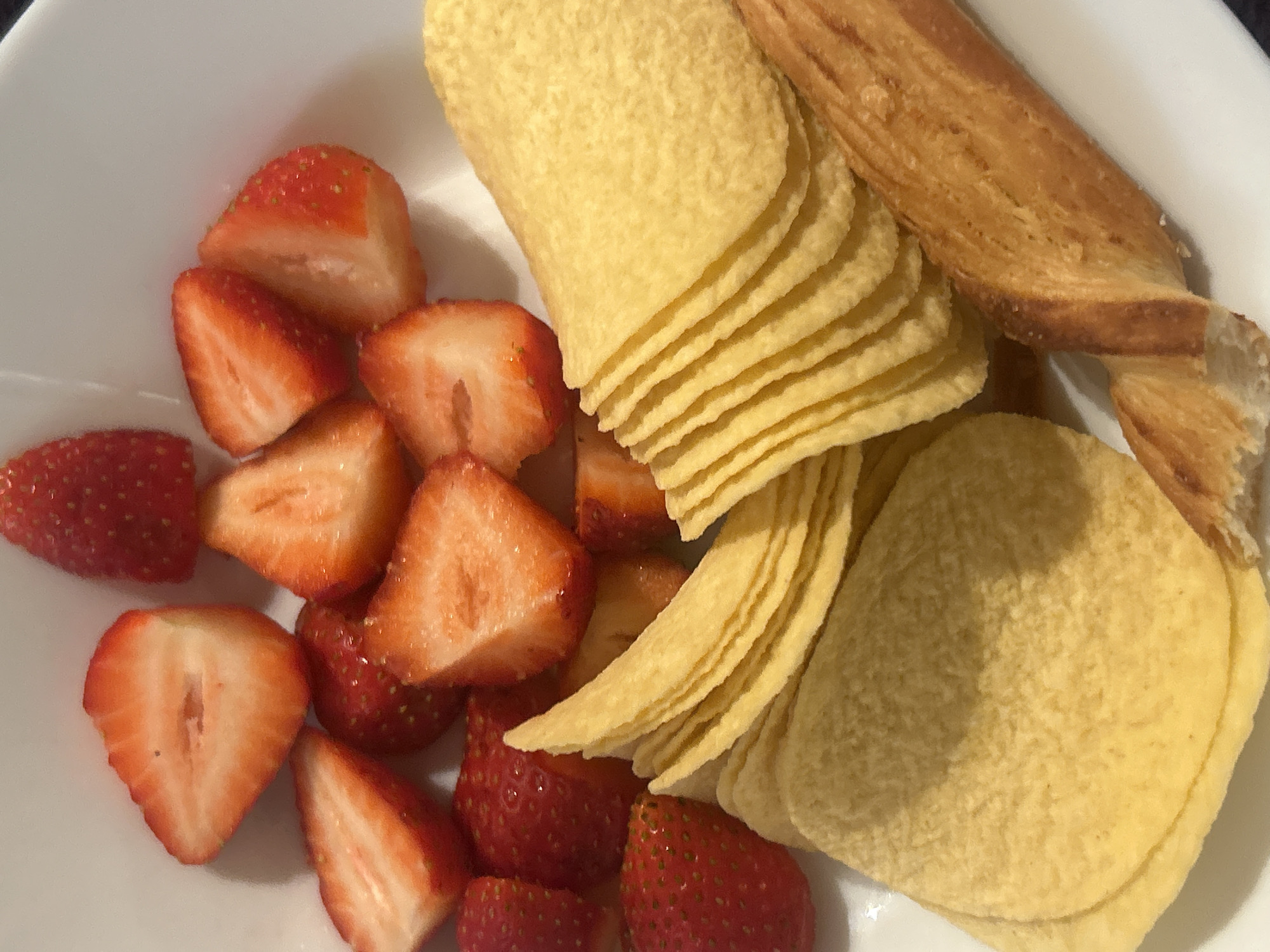 A white bowl contains sliced strawberries, a stack of potato chips, and a twisted pastry. The bowl is located in an unknown city.