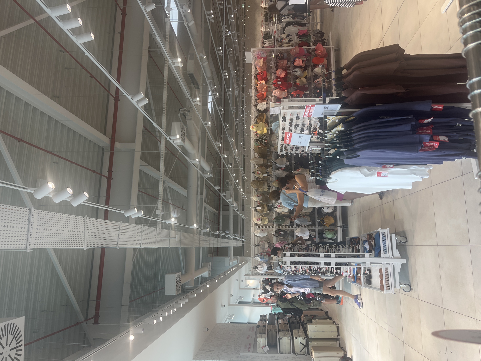The image depicts the interior of a retail store, likely a department store or a large clothing and accessories shop, located in Burgas, Bulgaria. The environment is brightly lit with a high ceiling featuring exposed industrial ductwork and numerous track lights. The store displays a variety of merchandise, including suitcases, hats, bags, sunglasses, and clothing items such as t-shirts and dresses, arranged on shelves, racks, and display stands. Several shoppers are visible browsing the merchandise, with some interacting with the displays. The flooring appears to be tiled. The overall impression is one of a busy retail space.