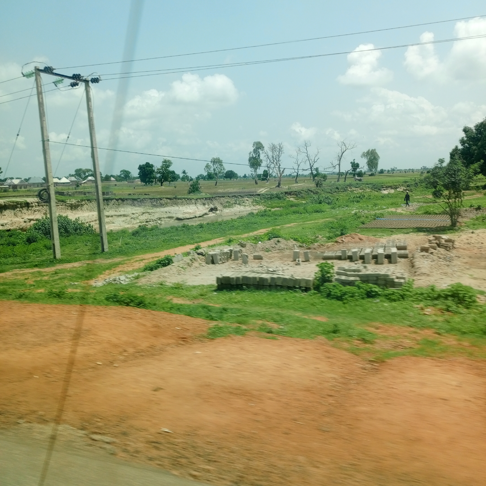 The image captures a rural landscape in Mutum Biyu, Nigeria, characterized by a light blue sky with scattered white clouds. The foreground is dominated by an orange-brown dirt road, blurred by motion, which transitions into a grassy area. Beyond the grass, a shallow, murky watercourse is visible, bordered by eroded, sandy banks. Construction materials, specifically stacks of concrete blocks, are present in a cleared area on the right side of the watercourse. Further in the background, several residential buildings with light-colored roofs are situated amidst a scattering of green trees and vegetation. A power line supported by two wooden poles runs horizontally across the mid-ground, with thick black wires extending from them. A single figure, dressed in dark clothing, is walking in the distance on the right side of the frame, near a flat, patterned surface that appears to be drying bricks or tiles. The overall scene depicts a developing rural area with ongoing construction and basic infrastructure.