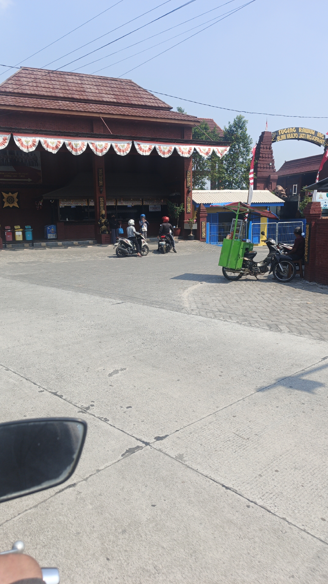 In Randugenengan, Indonesia, a street scene unfolds under a clear blue sky. A traditional brick building with a red tiled roof and decorative banners is visible on the left, with an entrance area featuring various stalls or shops. Several people are present, some on motorcycles. Two individuals are seated on a motorcycle on the right, parked next to a cart with a green cooler box. In the center, two motorcycles are parked, each with a rider wearing a helmet. Another motorcycle is parked further to the left. To the right of the main building, a gateway structure with Indonesian flags is prominent, and a small blue and yellow building is also visible. The foreground shows a concrete and paved road, with a motorcycle's side mirror and handlebar visible in the lower left corner, suggesting the image was taken from the perspective of someone riding a motorcycle. The overall impression is of a bustling, everyday scene in a local Indonesian setting.