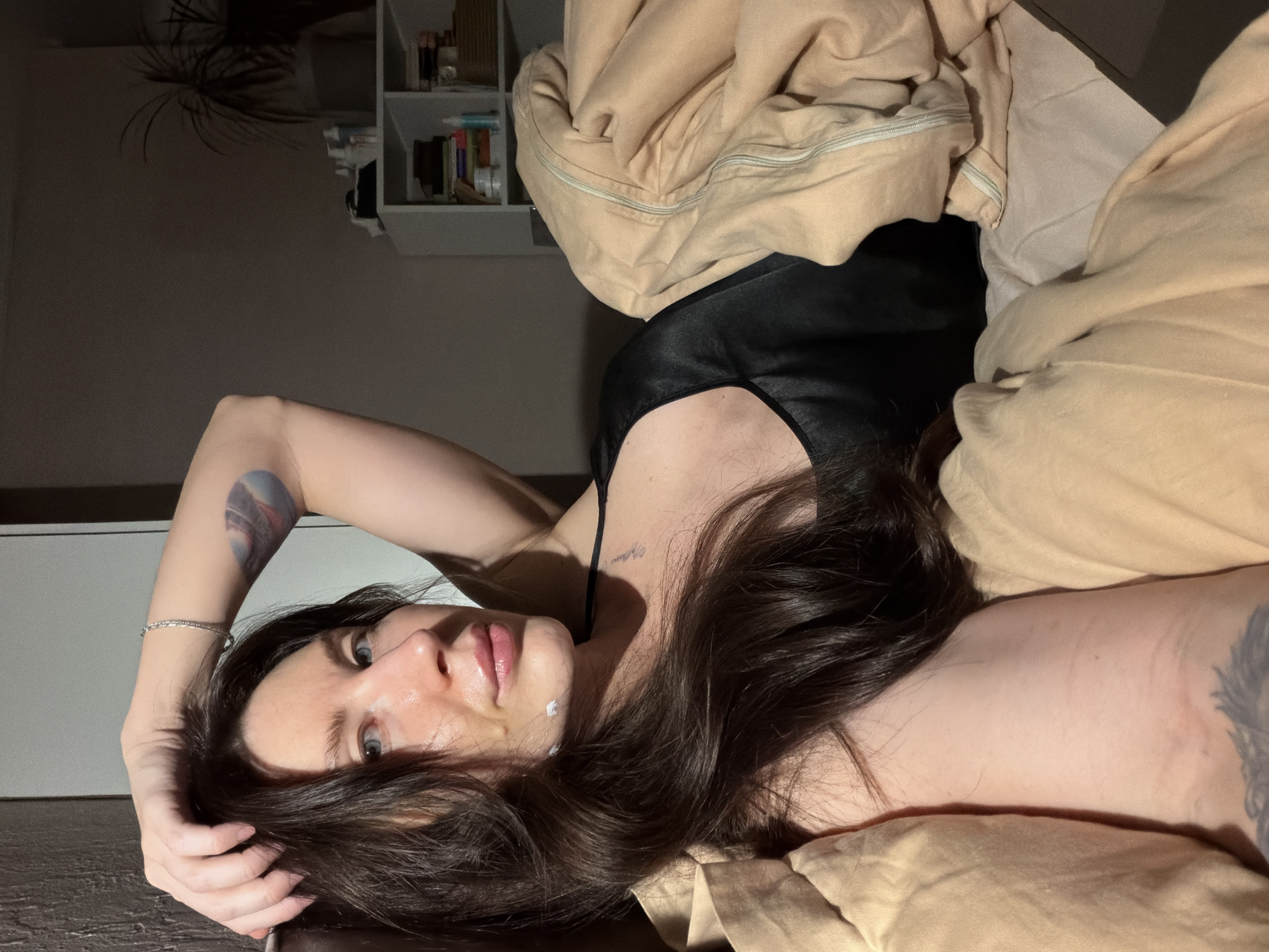 The scene depicts an adult female positioned in a bed within an indoor residential environment, specifically identified as being in Perm, Russia. The individual, appearing to be in her 20s or 30s, has long, dark hair and blue eyes. She is lying on her back, angled towards the camera, with her left arm bent and her hand resting on the back of her head, lightly touching her hair. A silver-colored braided bracelet is visible on her left wrist. On her left upper arm, a circular tattoo featuring abstract or celestial imagery in blue and pink tones is visible. She wears a black top, possibly a camisole or sleepwear. Two small white spots, consistent with a topical cream or product, are visible on her chin.
She is partially covered by light beige or tan bedding, specifically a duvet or comforter, which appears rumpled. The room is dimly lit, suggesting evening or nighttime. In the background, to the right of the subject, a white shelving unit is visible. This unit contains various items, including multiple bottles, what appears to be books or stacked papers, and a woven basket. To the far right, behind the shelving unit, a potted plant with dark green foliage is present. The walls are a neutral, dark gray color.