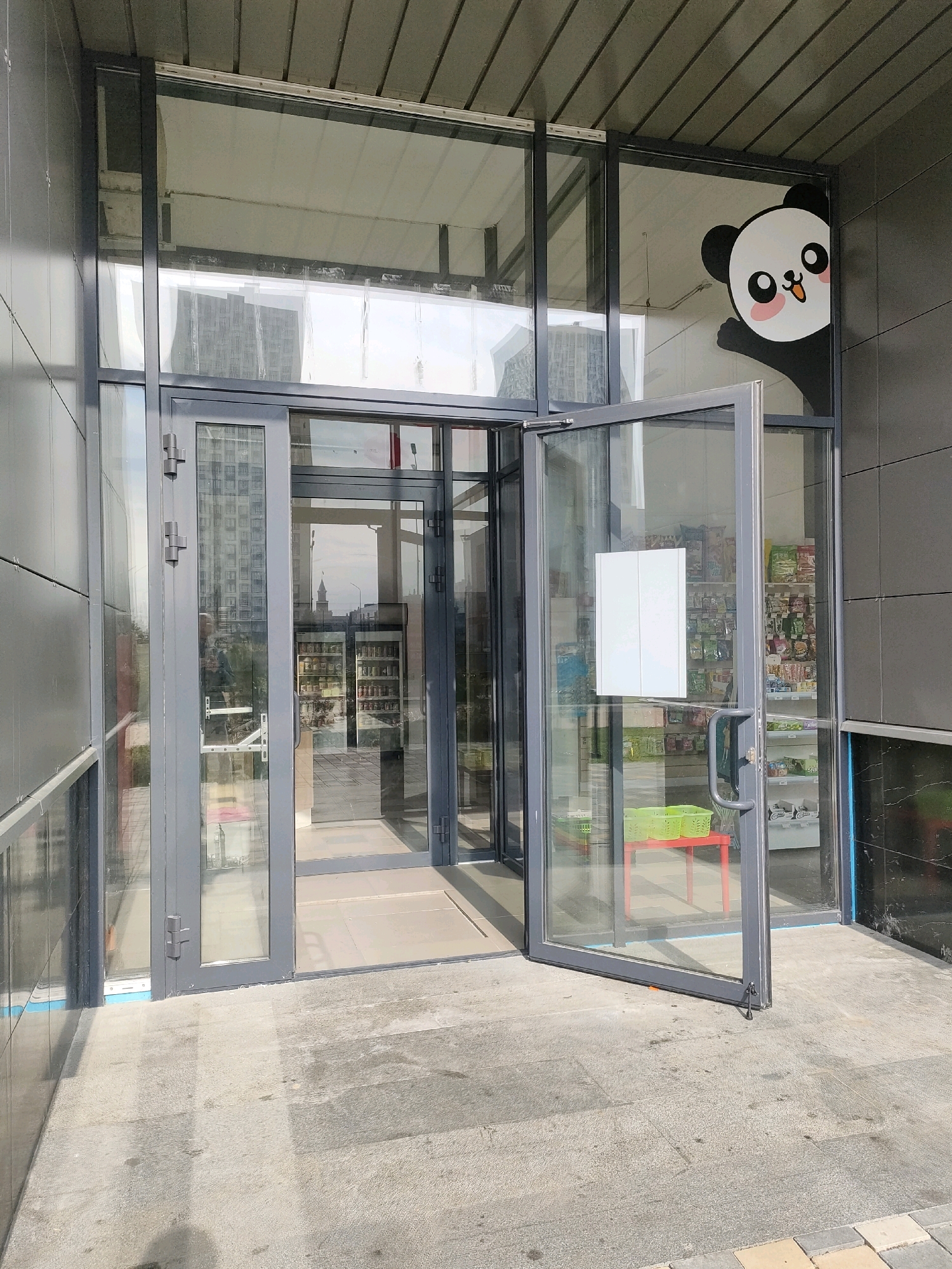 The image shows the entrance to a store with a glass facade. An open glass door reveals shelves stocked with packaged goods. A cartoon panda sticker is affixed to the upper right corner of the glass. The store is situated in Yekaterinburg, Russia, as indicated by the Cyrillic lettering visible on the store's signage within the image. The exterior is paved with grey flagstones, and the building's exterior walls are dark grey.