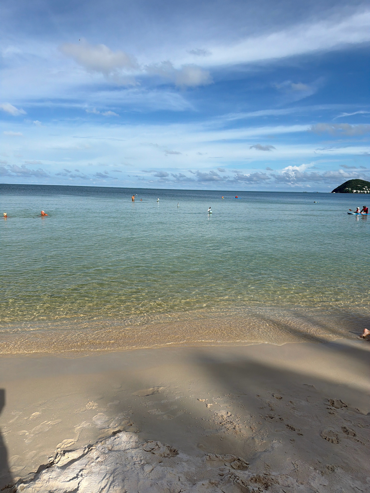 This image depicts a tranquil beach scene on the island of Phú Quốc, Vietnam, likely on the western coast, known for its calm waters and soft sands. The foreground is dominated by a sandy beach with visible footprints and a shadow cast across it, suggesting it's either morning or late afternoon. The water is remarkably clear, with gentle waves lapping at the shore, revealing the sandy bottom beneath. Several buoys are scattered in the shallow water, marking a swimming or recreational area. In the middle ground, a few individuals are enjoying the water: one person appears to be swimming, while others are on a paddleboard. Further out, a small, tree-covered island with white buildings is visible on the horizon to the right, a common sight along Phú Quốc's coastline. The sky above is a vibrant blue, dotted with white, fluffy clouds, indicating fair weather. The overall impression is one of a peaceful, sunny day at a tropical beach resort.