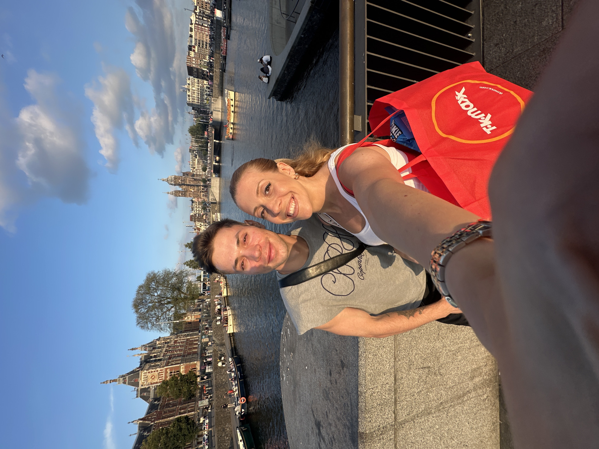 A selfie is taken by a couple standing on a bridge in Amsterdam, Netherlands, with a canal and historic buildings in the background. The man on the left is wearing a grey sleeveless t-shirt with a graphic print, and the woman on the right is wearing a white tank top. The woman is holding a large red TK Maxx shopping bag, which contains a blue bag of what appears to be crisps. In the background, a canal is visible with several boats docked and a few people seated on a stone ledge. The architecture suggests a historic European city, likely Amsterdam due to the distinctive canal system and building styles. The sky is blue with scattered white clouds, and the sun is casting a warm glow on the scene, indicating it is late afternoon or early evening.