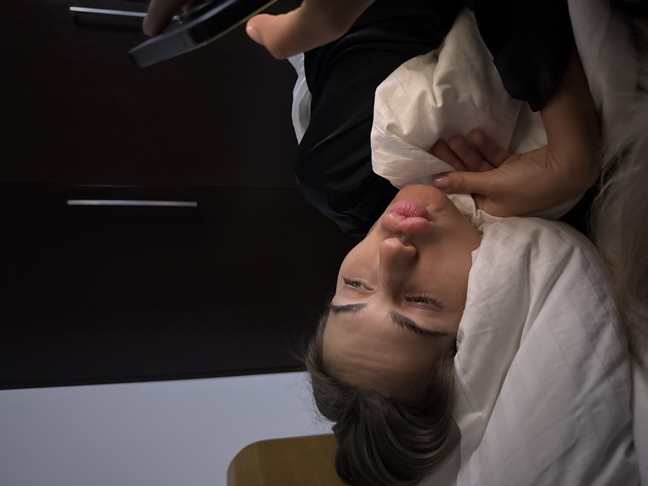 A woman with pursed lips lies in a bed covered by white sheets and a pillow. A hand holding a phone is positioned above, implying a selfie or video is being taken. Part of a dark wooden dresser is visible in the background, with a white wall beneath it. The image is slightly rotated, giving a tilted perspective. The woman is looking towards the camera with one eye slightly squinting.