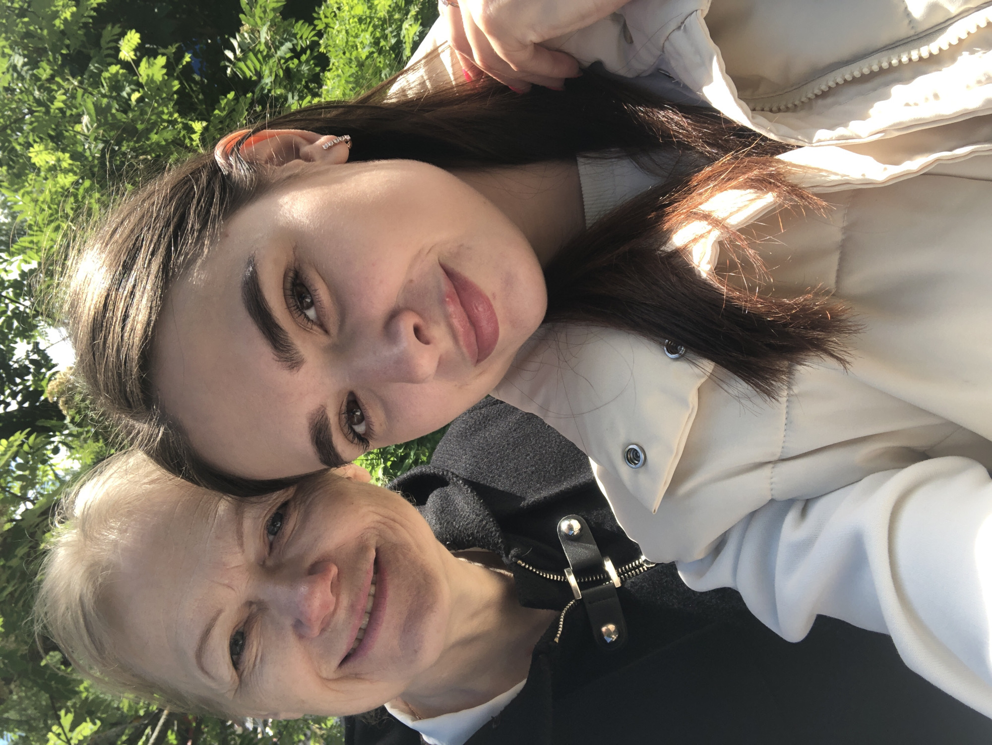 Two women are closely positioned in the frame, appearing to be taking a selfie. The younger woman has long brown hair, fair skin, and is wearing a light-colored padded jacket. Her face is tilted towards the camera, and she is smiling softly. The older woman has short, light-colored hair and is also smiling. She is wearing a dark-colored garment with a zipper and silver hardware.
Green foliage is visible in the background.
Based on the style of dress and general appearance, the photo could be taken in a public outdoor space. The location of Chaikovsky, Russia, is not visibly identifiable within the image.