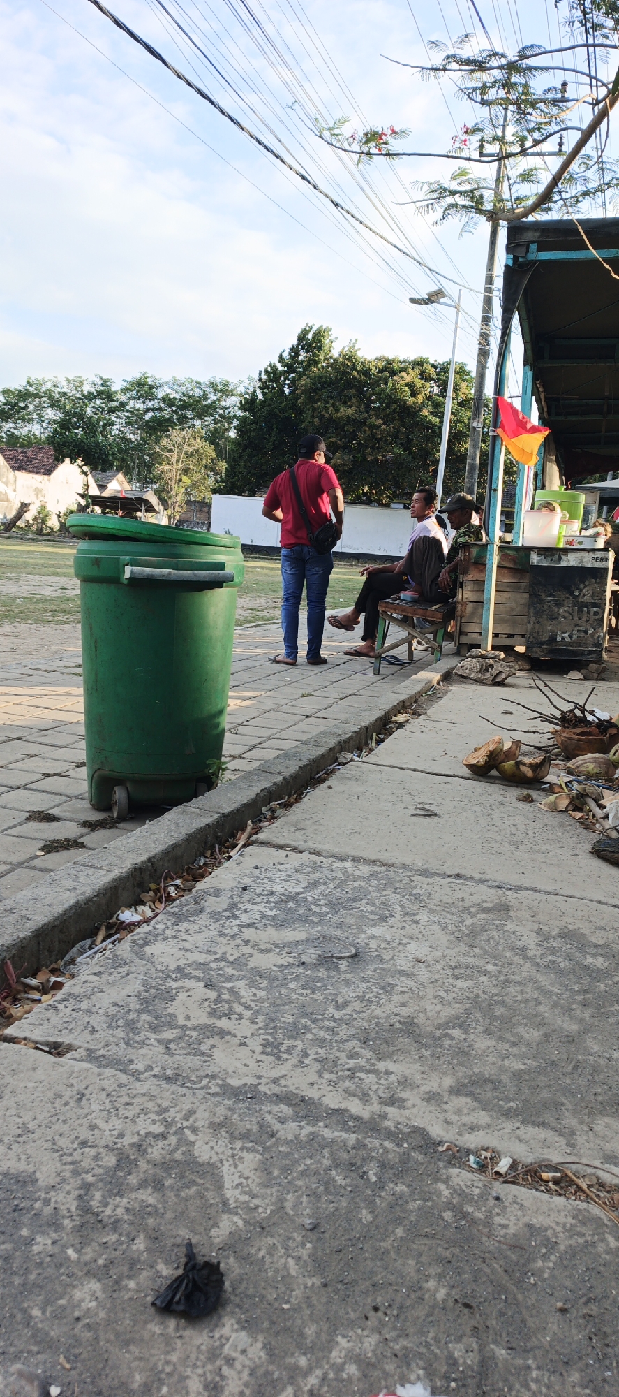 A full-shot, vertical, outdoor, daytime, candid photograph captures a scene on a paved area in Kasiyan Timur, Indonesia. In the foreground, a large green trash bin with a lid slightly ajar stands on a tiled walkway. The walkway is bordered by a concrete curb filled with debris. To the right, a concrete path shows scattered pieces of coconut husks and other organic matter. In the background, three men are present. One man in a red shirt and blue jeans stands facing away from the camera, with a black sling bag across his chest. He appears to be talking to another man sitting on a bench to his right. This second man is wearing a white shirt and dark pants, and is also facing away from the camera, in conversation with a third man who is sitting to his right, wearing a green shirt and a dark cap. This third man is partially obscured and also facing away. Behind the men, a wooden structure, possibly a stall or shop, is visible with a colorful flag attached to its roof. The overall environment suggests a public outdoor space, possibly near a marketplace or community area. The sky is partly cloudy, and trees are visible in the distance, along with some buildings.