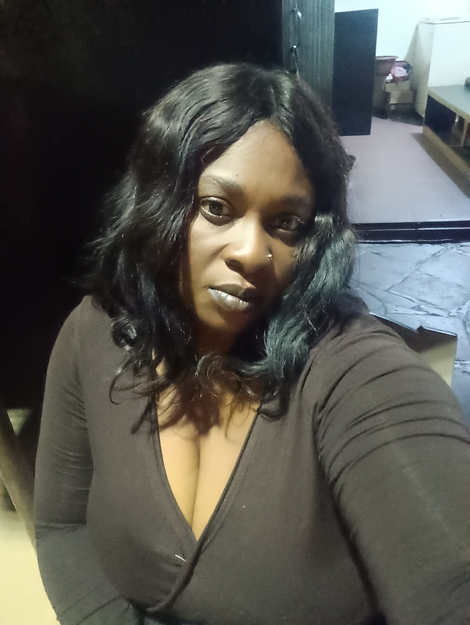 A medium-shot photograph depicts a woman with dark hair and brown skin. She wears a brown long-sleeved, low-cut top. The woman is the primary subject. Her face is visible, and she has a nose piercing on her left nostril. The setting appears to be an indoor location, possibly a residence in the city of Jos, Nigeria, as specified in the prompt. Details include a dark-colored piece of furniture in the background, along with a glimpse of another room with light-colored walls. The image quality is low, and the lighting is dim.