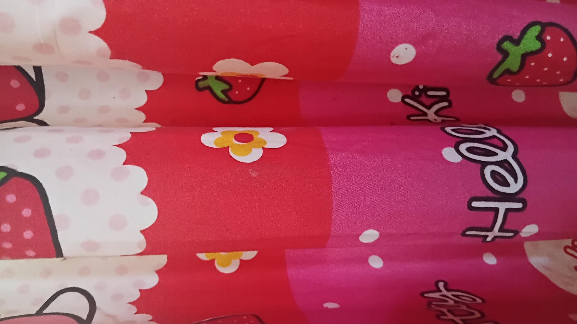 The image shows a close-up view of a pink and red patterned fabric, likely a curtain or shower curtain, featuring cartoon strawberries and flowers. The fabric has a playful, childlike design with white polka dots scattered throughout. Some sections of the fabric also have the words "ty" and "Hello Ki" printed on them in white script. The scene is indoors and appears to be in a domestic setting. There are no people or specific city landmarks visible to determine a location within Prabumulih, Indonesia. The lighting suggests an indoor environment.