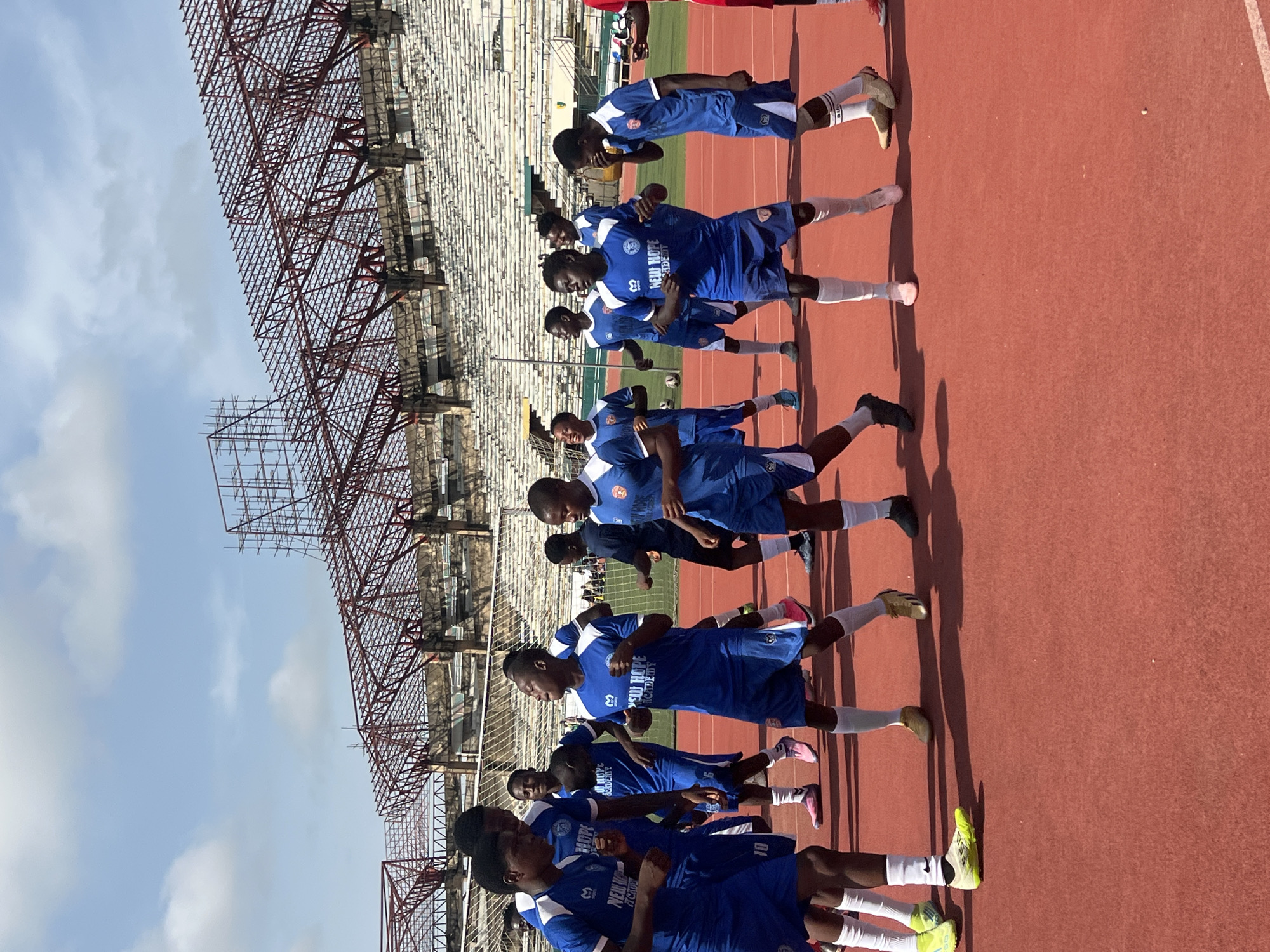 In the city of New Stadium, Nigeria, a group of young male athletes in blue and white soccer uniforms are on a red track field, seemingly engaged in a warm-up or drill. The stadium's tiered seating is visible in the background, along with its metal framework, indicating ongoing construction or renovation. The sky is a clear blue with scattered clouds. The athletes' expressions range from focused to jovial, suggesting a practice session in good spirits. The scene captures a moment of athletic activity within a developing sporting venue.