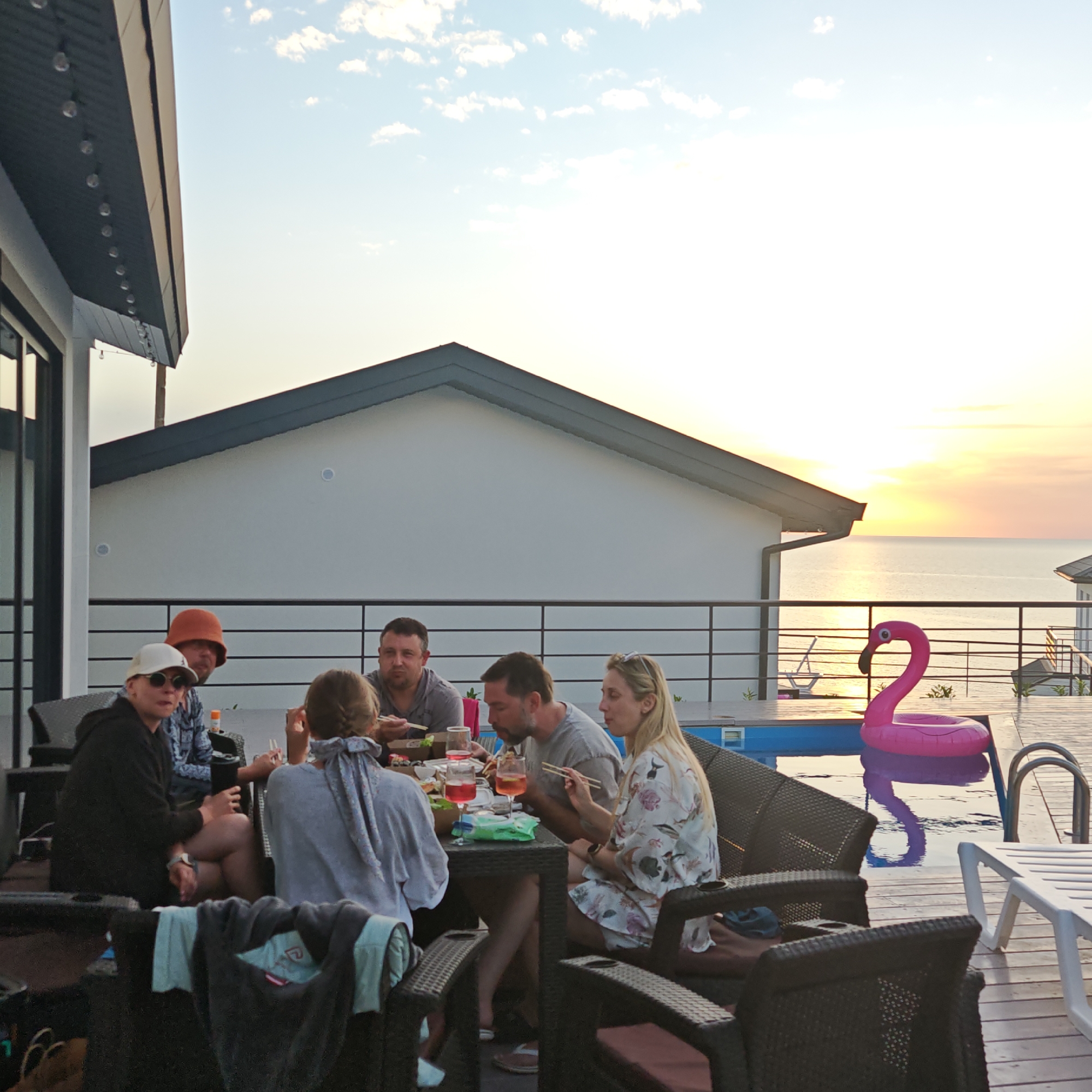 A group of six people are gathered around a table, seemingly enjoying a meal with chopsticks. The setting appears to be a patio or deck area overlooking a body of water, possibly an ocean, at sunset. The area includes a pool, with a pink flamingo float. The architecture consists of a modern building with a flat roof and dark trim. The sky is visible with some clouds and the sun setting in the distance.