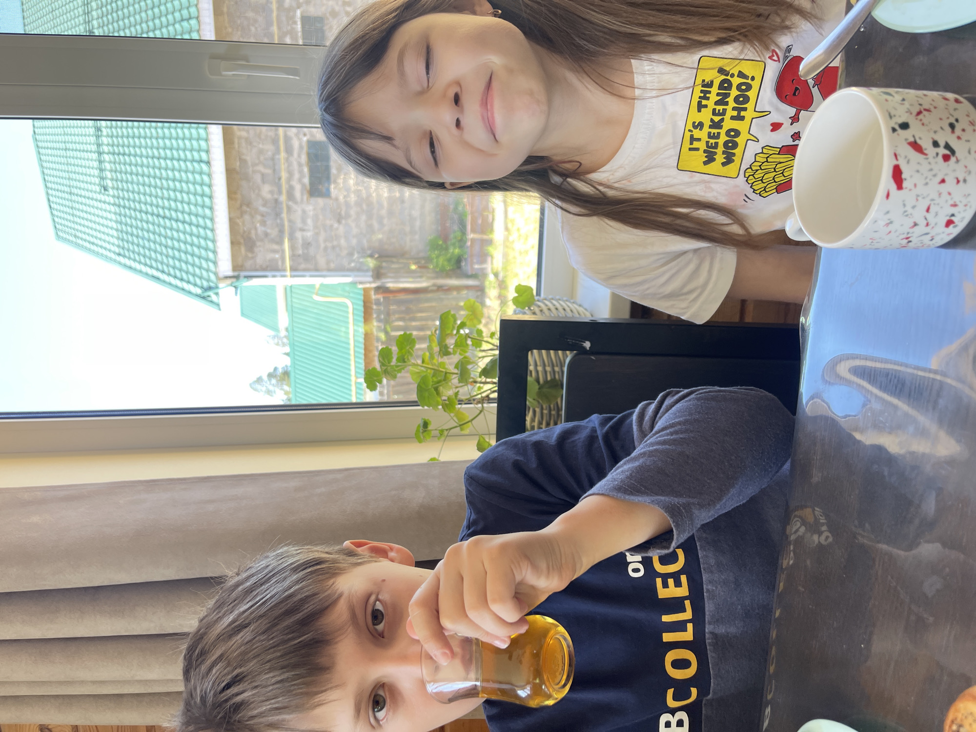 Two children are seated at a table indoors. A girl with long brown hair is in the upper part of the frame, smiling with her eyes closed. She is wearing a white t-shirt with a cartoon graphic. A white and red patterned mug sits on the table to her right. Below her, a boy is holding a glass of amber-colored liquid up to his mouth. He has short brown hair and is wearing a dark blue long-sleeved shirt with yellow lettering. A window is behind them, showing a green-roofed building and a plant. The scene is in Chaikovsky, Russia.