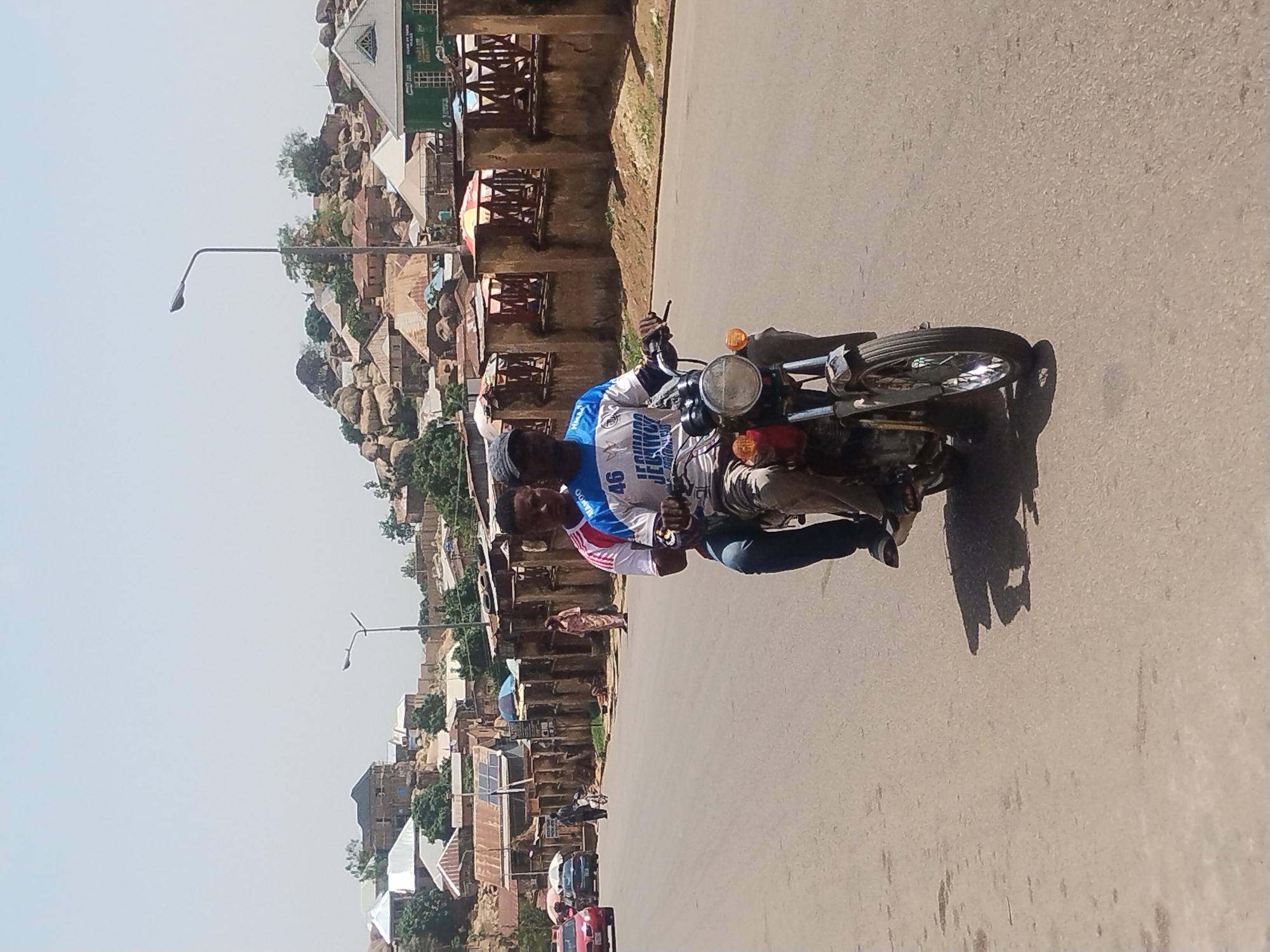 Two men ride a motorcycle on a paved road in Jos, Nigeria. The road is lined with buildings, many with corrugated metal roofs. One man rides the motorcycle, and the other man sits behind him. They are both wearing casual clothing. The city is situated in a hilly region. A lamppost is visible on the left side of the road. Several vehicles are driving on the road. A pedestrian walks on the sidewalk.
