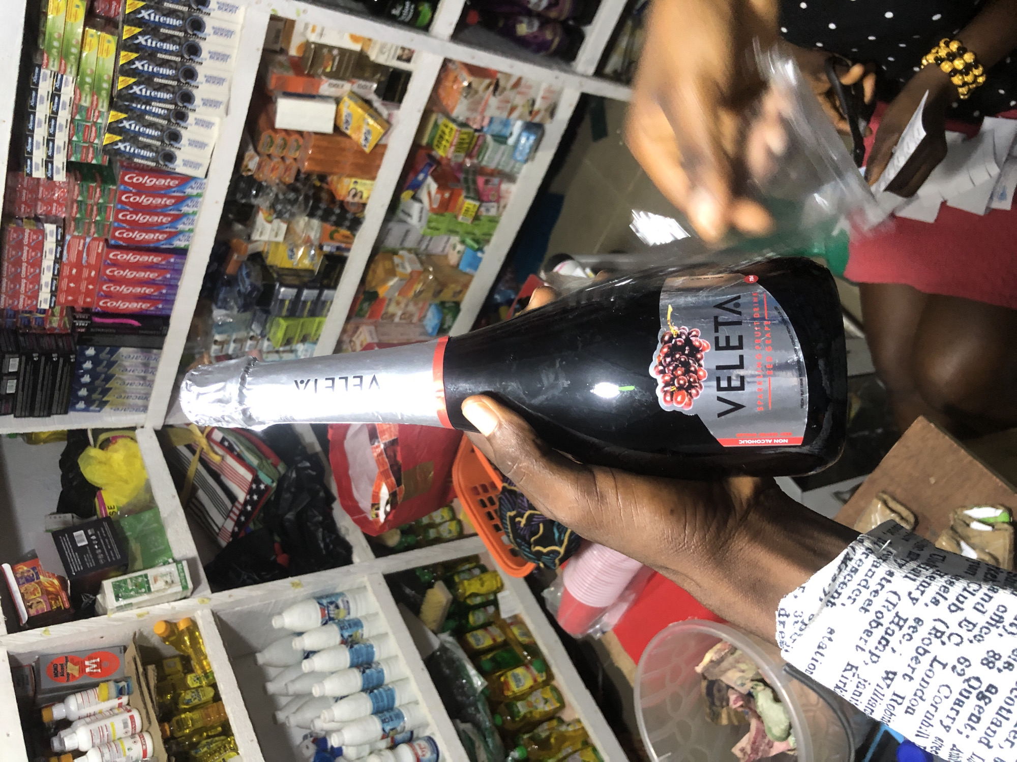 A dark-skinned person's left hand holds a bottle of Veleta sparkling fruit drink in the foreground, with the bottle partially obscuring a shelf stocked with various consumer goods, including bottles of cleaning supplies and colorful boxes of toothpaste. The background reveals a cluttered shop environment with shelves filled with merchandise. A red basket and a red plastic cup are visible on a lower shelf. The person is wearing a white shirt with a newspaper print. In the background, another person's hands are visible, holding scissors and a receipt. The scene appears to be a shop in Ado, Nigeria.