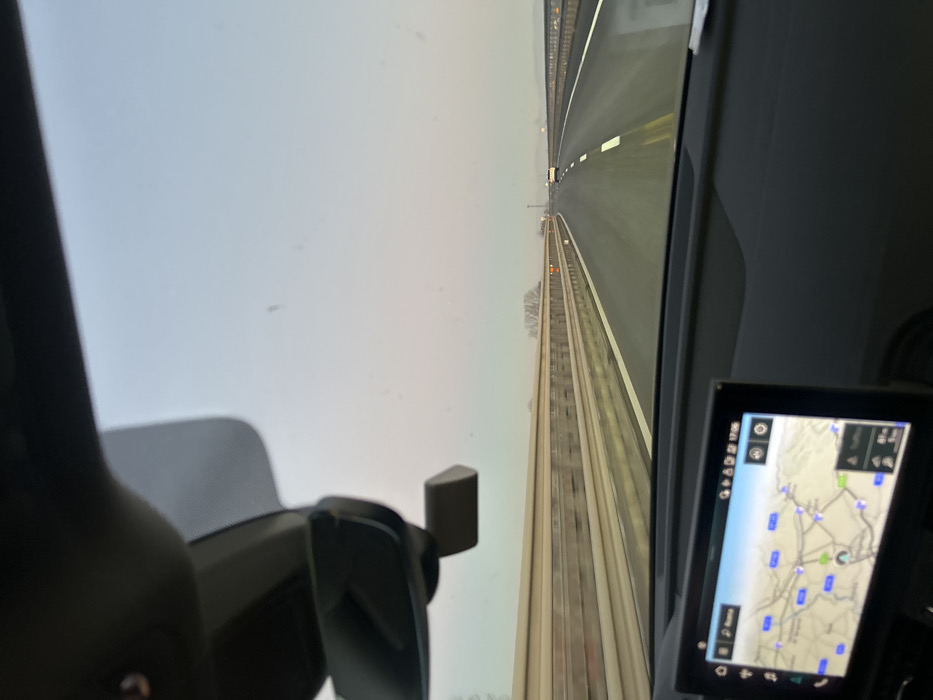 The image, oriented vertically, depicts a view from inside a moving vehicle looking forward onto a railway track. The track, composed of rails and ties, extends into the distance under an overcast or hazy sky. Parallel to the main track on its right side, another track or paved surface is visible, also stretching into the horizon. The muted lighting suggests either an overcast day or late afternoon/early morning conditions.
In the bottom left of the frame, a dark grey interior component, possibly part of a seat or dashboard, and a black mounting bracket (resembling a phone holder) are partially visible.
To the bottom right, a digital screen displays a navigation map. The map shows a network of roads, marked with blue rectangular location pins and a few green circular markers. The time "17:35" is visible in the top right corner of the screen, along with small icons for settings, volume, traffic, and battery/signal status. Based on the provided context, the map is displaying the region around Alessandria, Italy. The scene captures the experience of traveling along a railway line, likely from the cab of a train.