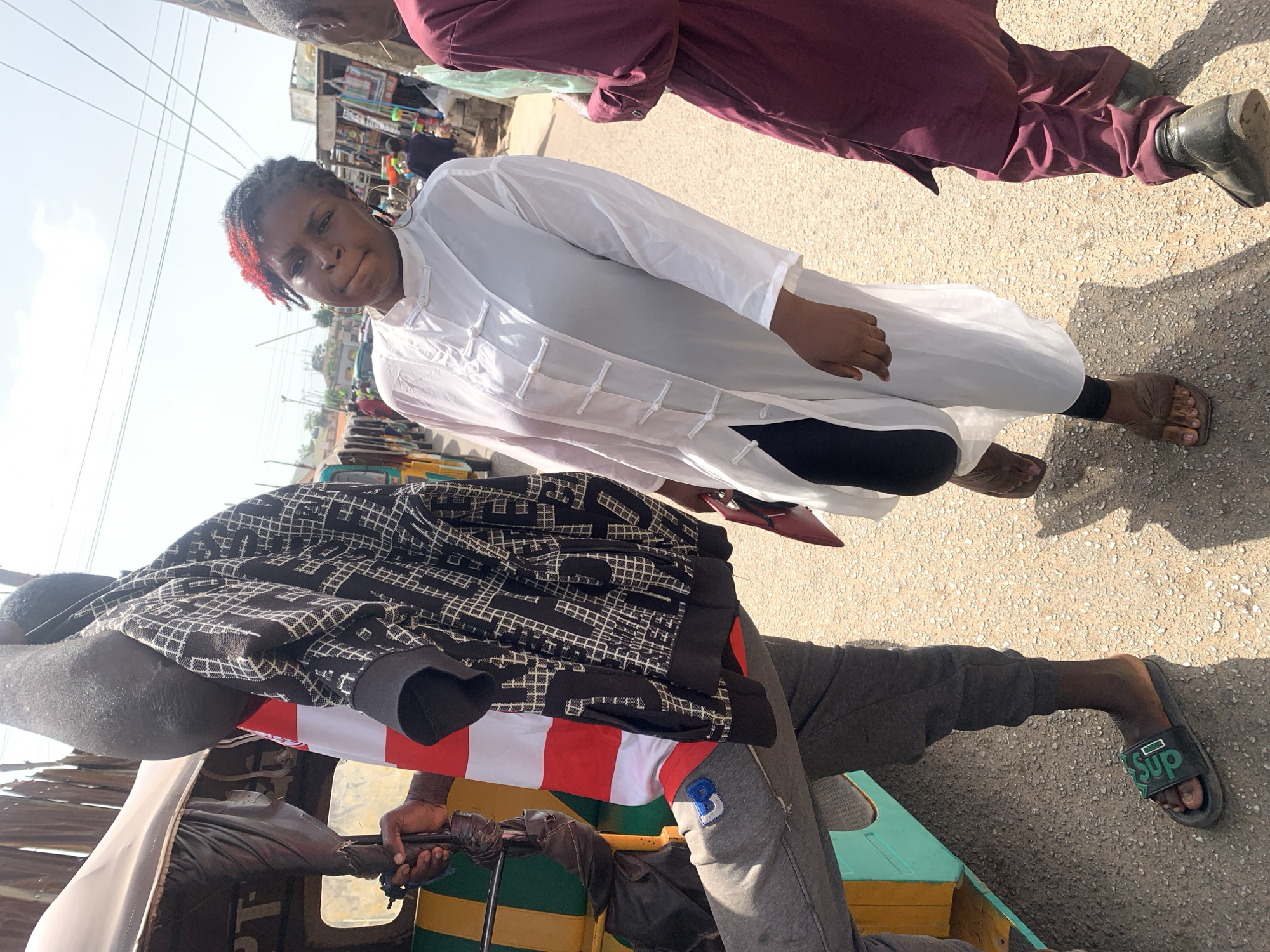 The image shows a woman in a white tunic-style shirt with black pants and sandals standing on a paved street, likely in Jos, Nigeria. Her hair is partially dyed red. She is flanked by other individuals; one in a maroon outfit with leather shoes and another in a black and white patterned jacket, gray pants, and green sandals sitting inside what appears to be a vehicle, possibly a tuk-tuk or tricycle common for transportation in the area. The street has other pedestrians and shops visible in the background, under a bright sky. Electrical wires are strung overhead.