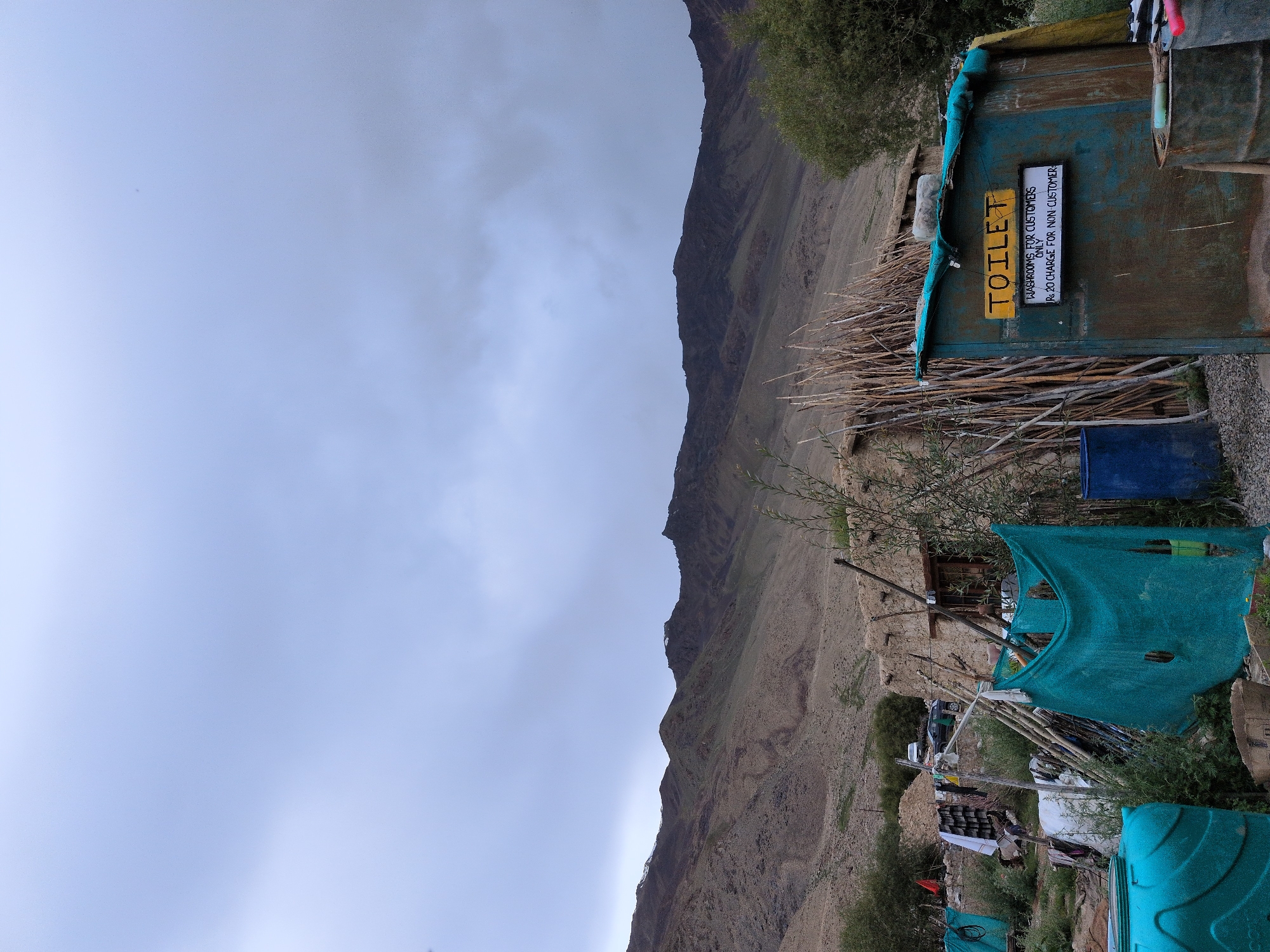 A rustic, weather-beaten toilet structure stands in a remote, mountainous area, possibly near Khardung, India. The structure is painted a faded teal and features a yellow sign that reads "TOILET". Below this, a smaller white sign states "WASHROOMS FOR CUSTOMERS ONLY Rs. 20 CHARGE FOR NON CUSTOMERS". The toilet is surrounded by a natural, arid landscape with rocky, barren hills in the background. Debris and makeshift structures, including poles and tarps, are scattered around the scene, suggesting a basic, temporary settlement or service area. A large blue plastic tank is visible to the left of the toilet, and a dark blue barrel is to its left. The sky is overcast with heavy clouds.