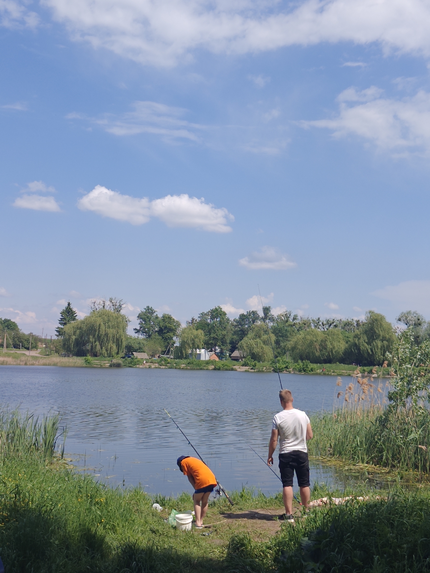 The photograph shows two people fishing on the shore of a body of water on a sunny day. One person, wearing an orange shirt and a blue hat, is bent over, possibly tending to fishing equipment. The second person, wearing a white shirt and dark shorts, stands upright with their back to the camera, also holding a fishing rod. The shoreline is grassy with reeds and other vegetation, while the opposite shore has trees and some visible structures in the distance, suggesting a rural or semi-rural setting. The sky is blue with scattered clouds. The location is likely Bobrivnyk, Ukraine, given the prompt's instructions.