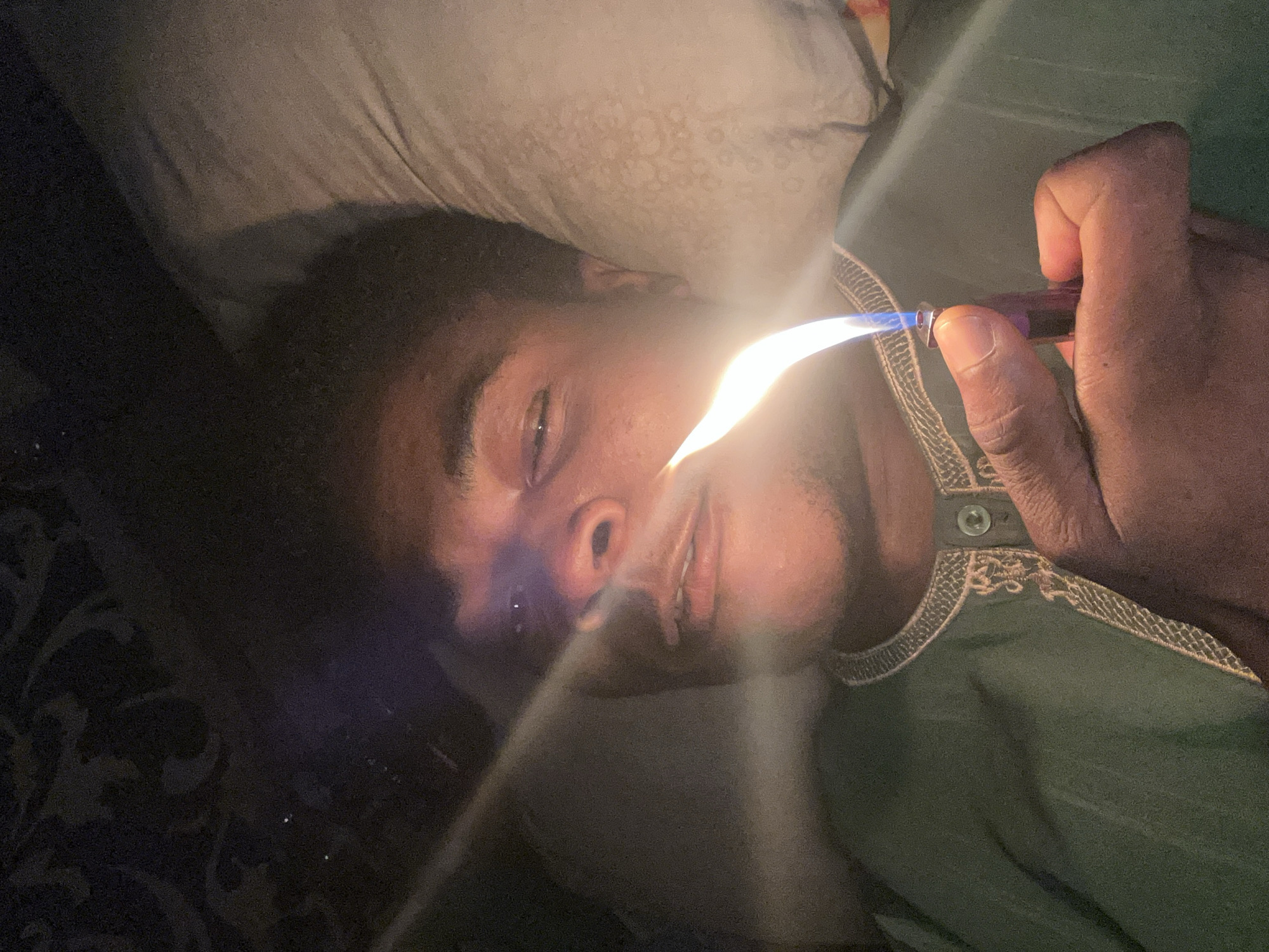 In this close-up, dimly lit image, a person is lying down, with their head resting on a pillow. A lighter is held in their right hand, with the flame igniting and producing a bright light that illuminates the person's face and the surrounding area. The person appears to be looking towards the light source with their eyes partially open. They are wearing a green garment with decorative trim and a button. The background is dark and indistinct, with some subtle patterns visible in the lower left portion of the image. The overall scene suggests an intimate or private moment, possibly at night or in a dark room. There is no visible text in the image. The setting is Ihiagwa, Nigeria.