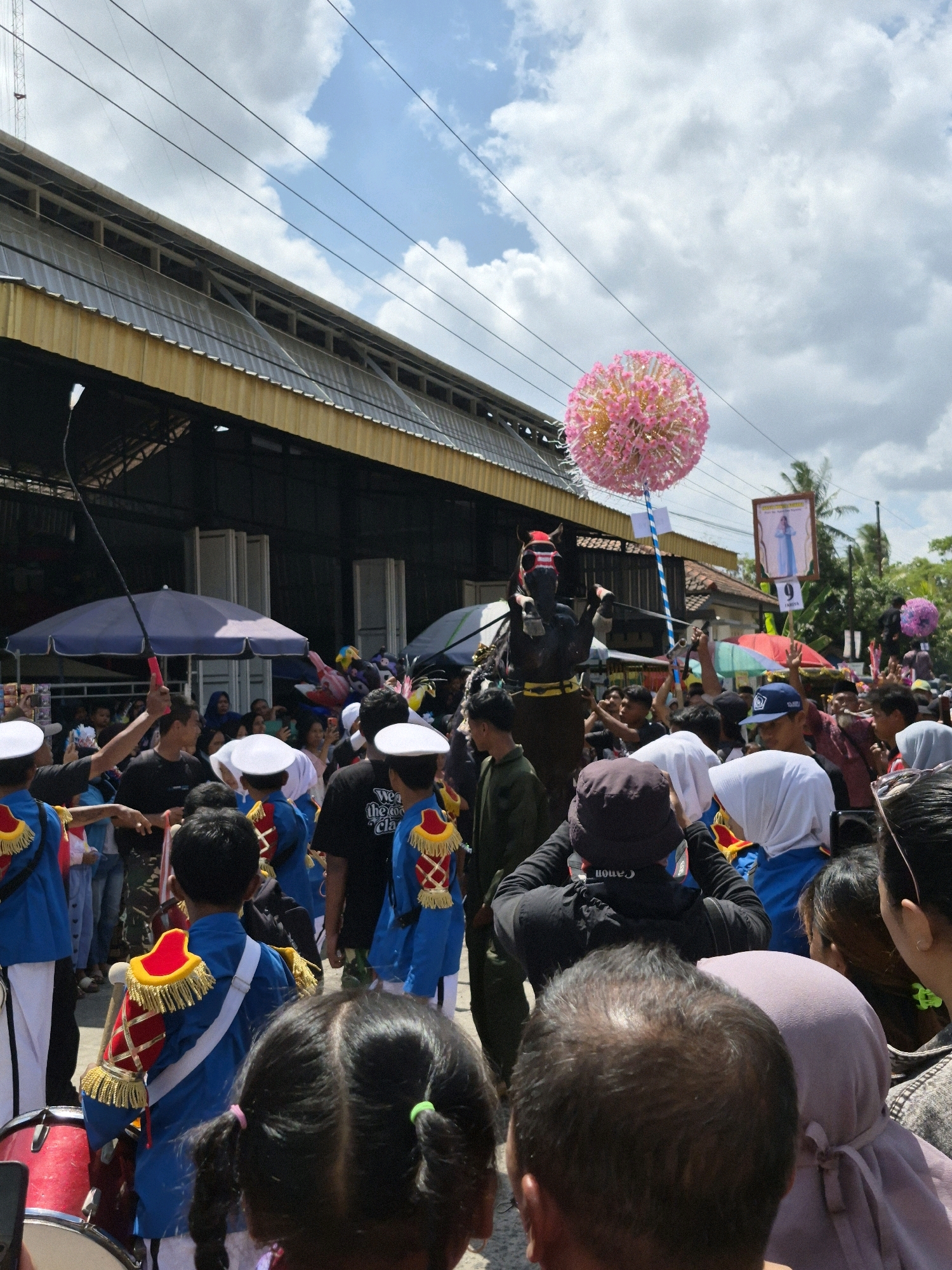 The image depicts a street scene in Grantung, Indonesia, during what appears to be a parade or festival. In the foreground, a young girl with dark hair tied in pigtails is seen from behind, looking towards a procession. Several children in blue marching band uniforms with red and gold trim are visible, some holding instruments. One child is wearing a white hat.
The procession includes a person on horseback, wearing a dark costume and a whip. Above the horse, a large, spherical pink decoration on a pole is held aloft. In the background, a crowd of spectators lines the street, some holding umbrellas and cameras. A building with a yellow awning stretches across the upper portion of the image, with power lines crisscrossing the sky. A framed image of a woman in blue is displayed on a pole in the background, suggesting a religious or cultural element to the event. The sky is partly cloudy.