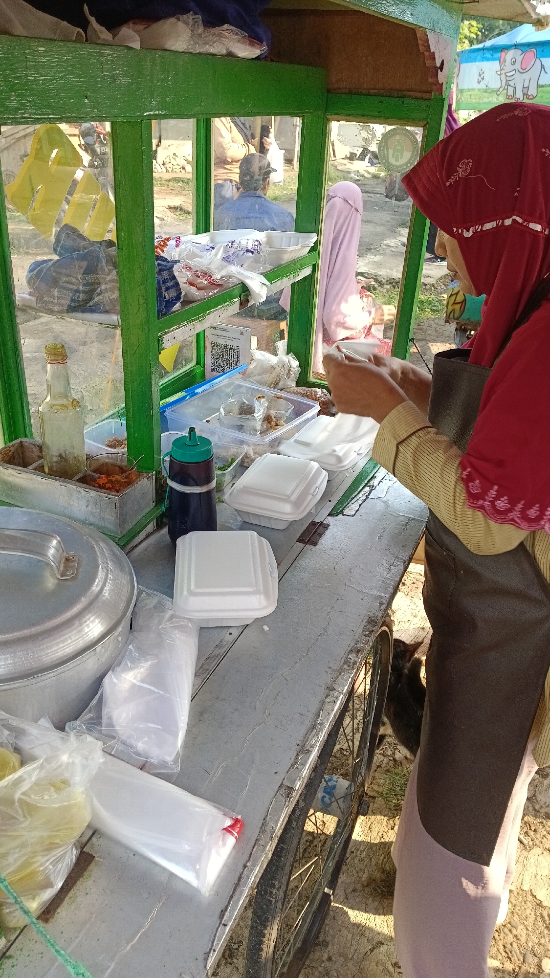 A street vendor in Tajurhalang, Indonesia is preparing food from a mobile cart. The cart is painted green and has glass panels. A woman wearing a red hijab and an apron is seen in the foreground, holding a white styrofoam container. She is interacting with a customer whose hands are visible. Various food containers, bottles, and plastic bags are on the cart's surface. In the background, other individuals are visible, and there are some buildings and a drawing of an elephant on the cart. The ground beneath the cart appears to be dirt and gravel. A black cat is partially visible near the cart's wheels.