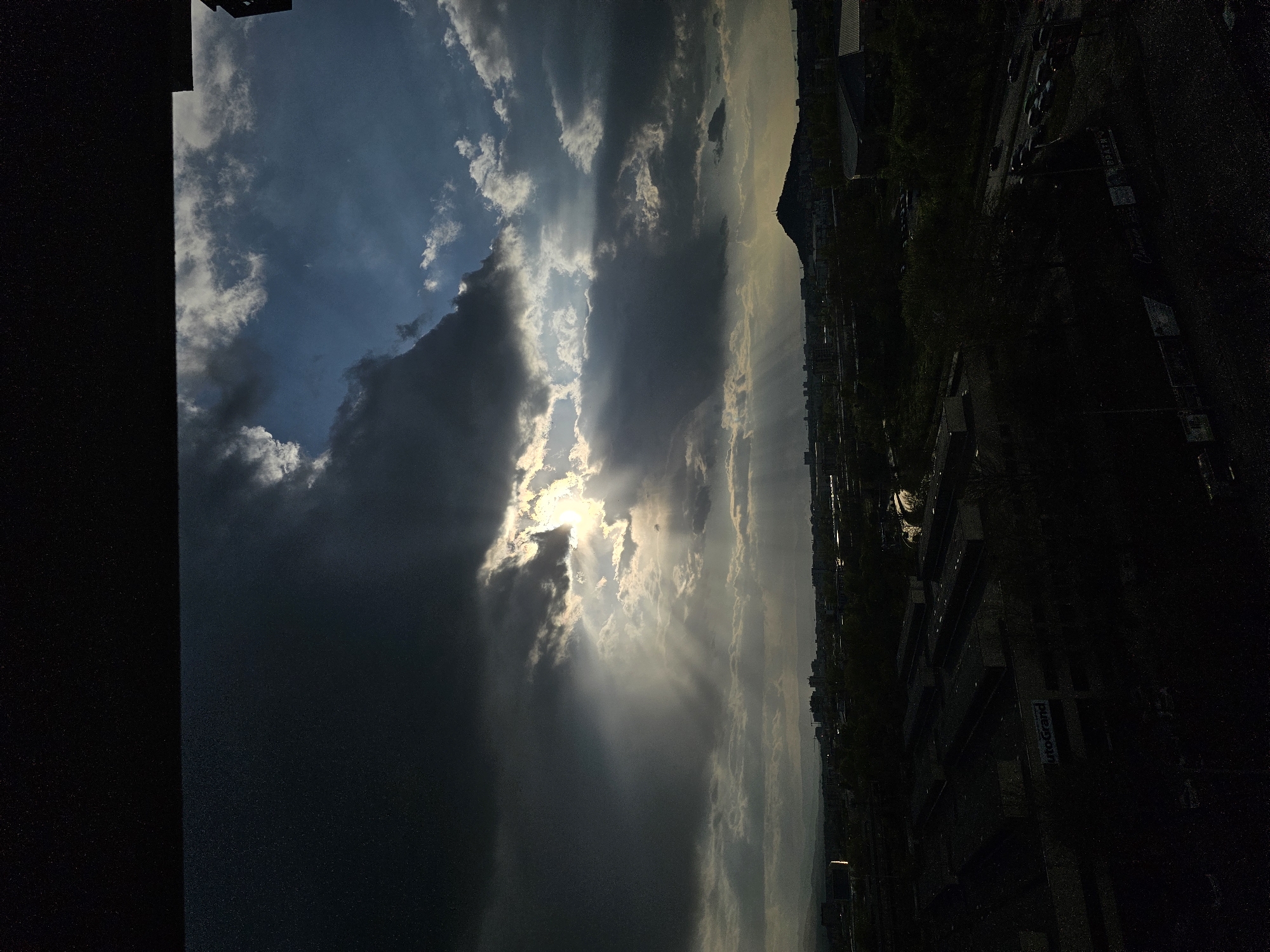 The image shows a view of the sky and a portion of Plovdiv, Bulgaria. The sky is dominated by dramatic clouds with the sun shining through, creating light rays. The city is visible on the right side of the image, appearing dark in contrast to the bright sky. Buildings, trees, and roads are discernible, but details are obscured due to the low lighting and viewing angle.