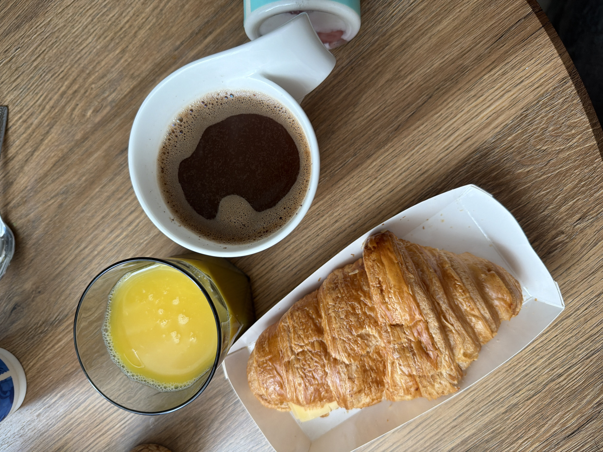 The image presents a top-down view of a meal setting on a wooden table. The central items are a cup of coffee, a glass of orange juice, and a croissant.
A white ceramic cup with a distinctive, bent handle shape holds a dark, frothy coffee with a layer of crema visible on its surface. To the lower left of the coffee, a clear glass is filled with bright orange juice, showing small bubbles. To the lower right of the coffee and beside the juice, a large, golden-brown croissant sits in a white, rectangular cardboard container. The croissant has a flaky texture and appears to be filled with a yellow ingredient, likely cheese.
The objects are placed on a wooden table with a noticeable grain pattern. Partial views of other items are visible: a section of a blue and white patterned object (possibly a coaster) is on the far left, and a blurred white and light blue container or lid is partially visible at the top edge of the frame, above the coffee cup.
The scene depicts a casual meal, most likely breakfast or a snack, occurring indoors during daylight hours. No people are visible. No discernible text is legible on any of the items. This scene is located in Khimki, Russia.
