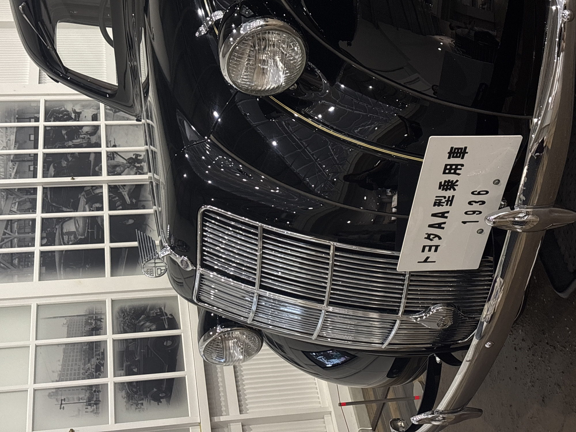 The image shows the front of a black vintage Toyota AA passenger car from 1936, displayed at the Toyota Commemorative Museum of Industry and Technology in Nagoya, Japan. The car features a prominent chrome grille, two round headlights, and a hood ornament. A license plate with Japanese text and "1936" is attached to the front bumper. Behind the car, a wall with large windows displays historical black and white photographs of early automobiles and factory scenes, offering a glimpse into automotive history. The environment appears to be an indoor exhibition space with controlled lighting, highlighting the car's polished surface.
