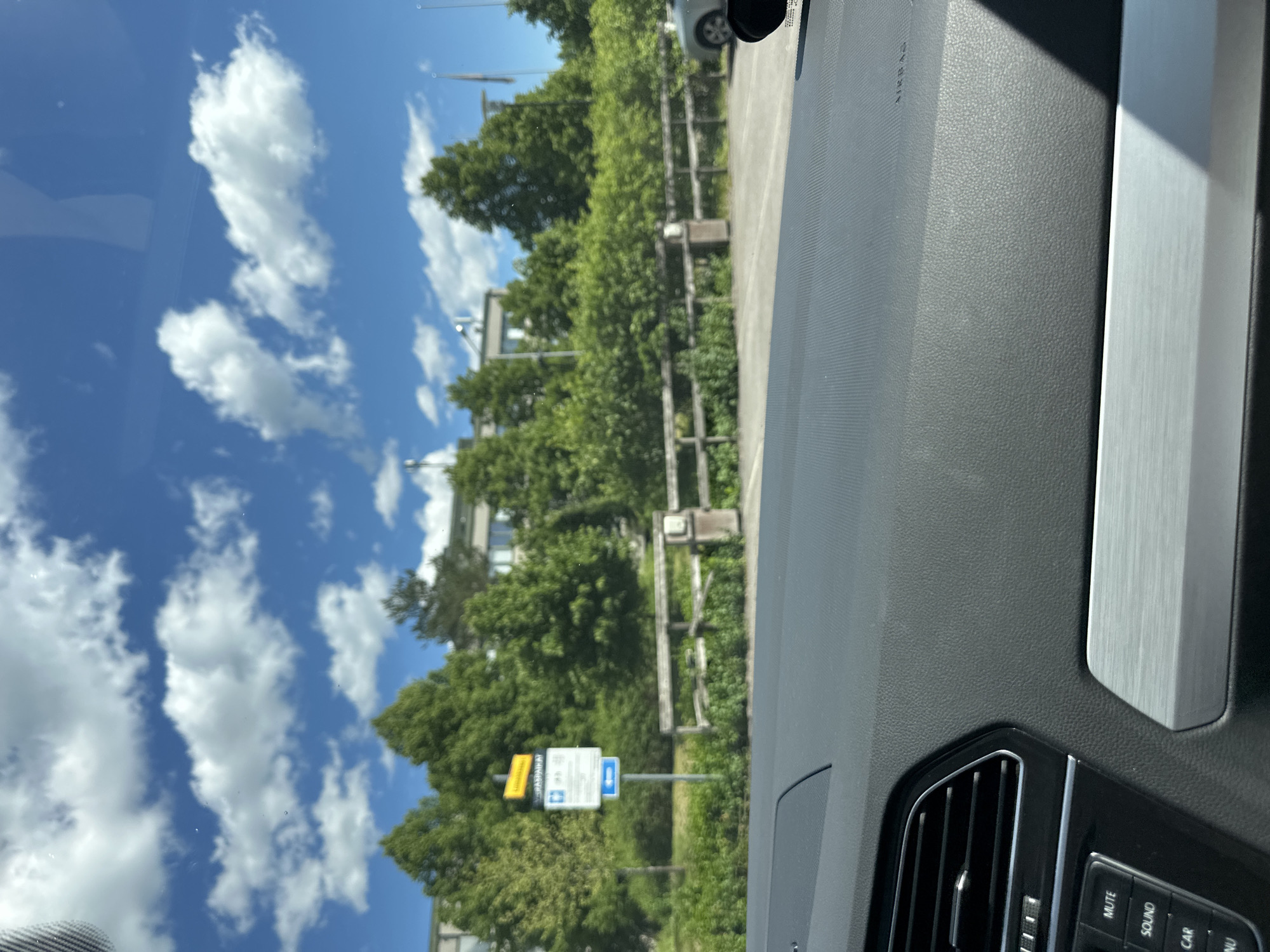 The image is taken from inside a vehicle, looking out through the windshield. The foreground shows the car's dashboard, with vents and buttons visible. Beyond the car, there is a parking area with a wooden fence and some bushes. In the background, a modern building is visible, surrounded by lush green trees under a blue sky with scattered white clouds. A street sign indicating "Pysäköinti" (Parking) and a blue arrow is also present on the left side of the image. A silver car is parked in the distance on the right. The scene appears to be a sunny day in an urban or suburban environment.