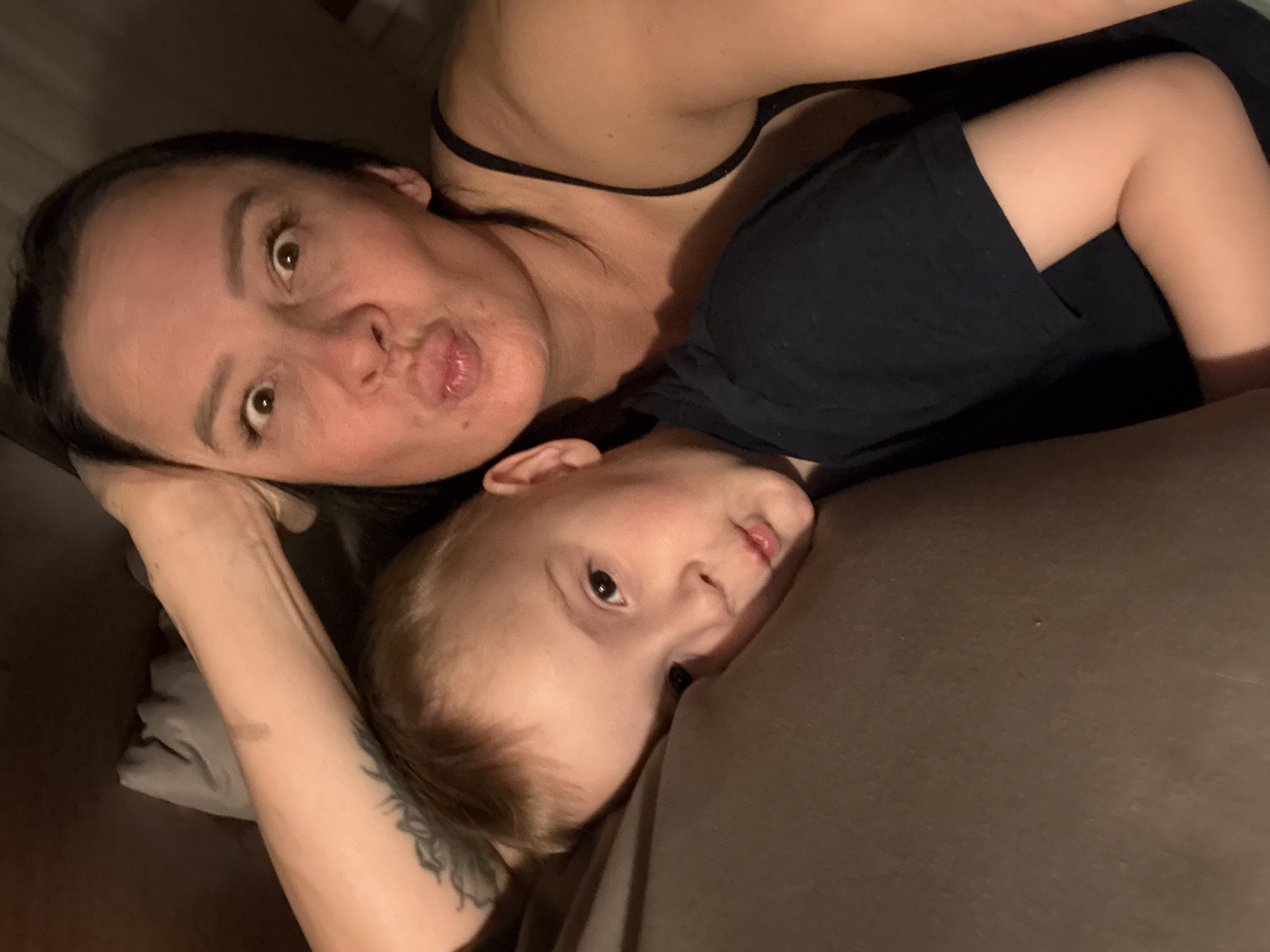 This is a close-up, intimate portrait featuring an adult female and a young child, likely a selfie taken in an indoor, dimly lit environment, possibly during the evening or night. The image is oriented vertically.
The adult female, positioned on the left, has dark hair pulled back and brown eyes. She is looking directly at the camera with a playful expression, her lips pursed in a "kiss" or "duck face." She is wearing a dark, thin-strapped top, and a tattoo with a detailed, possibly floral or abstract, design is visible on her left forearm. A minor abrasion or mark is also seen on her inner arm.
Resting closely beside her, on her arm or chest, is a young child with light brown hair and brown eyes. The child is also looking towards the camera with a curious, neutral expression, and has slightly parted lips. The child is wearing a dark, short-sleeved top, and one of their bent legs, also clad in dark fabric, is visible in the lower right portion of the frame.
Both individuals are lying on a soft surface, which appears to be a dark brownish-grey fabric, characteristic of a bed or sofa. The low light and close proximity suggest a moment of relaxation and affection. There are no specific location cues visible in the image to indicate Chaikovsky, Russia, nor is there any visible text.