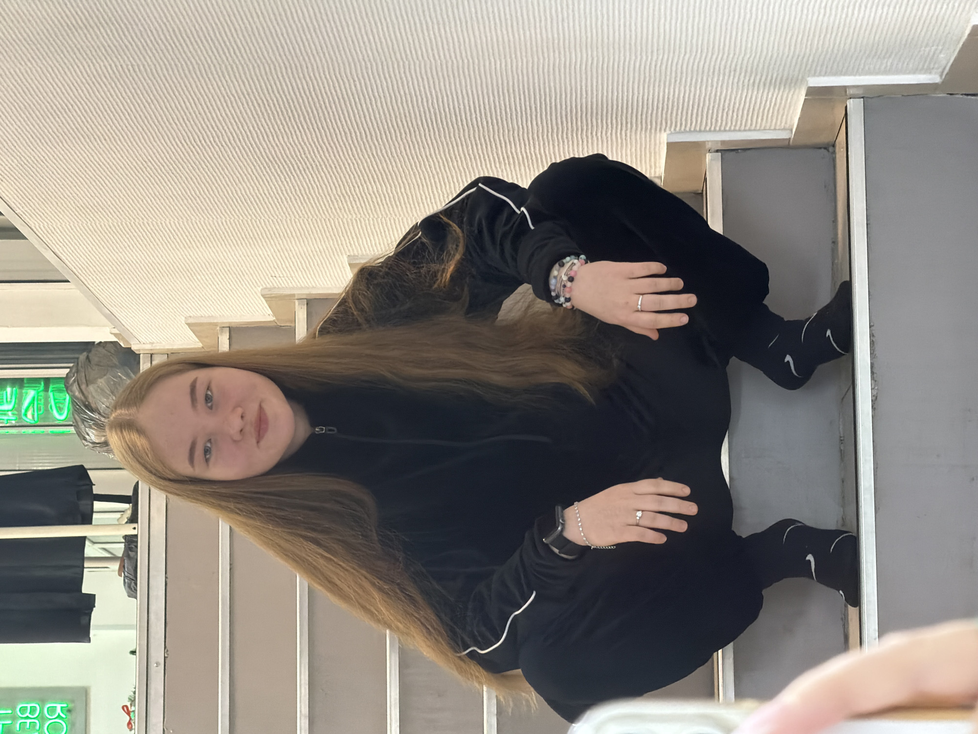 A young female is photographed squatting on an indoor staircase in the city of Chaikovsky, Russia. She possesses long, straight, light brown hair and gazes directly at the camera with a neutral expression. She is attired in a black velour tracksuit featuring white piping along the sleeves and black socks with white Nike swoosh logos. On her left wrist, she wears a dark-colored smartwatch and a silver-colored bracelet with small clear beads. A silver-colored ring with a single clear stone is present on her left ring finger. Her right wrist displays multiple colorful beaded bracelets, and her right ring finger has a plain silver band. The staircase steps are gray with lighter risers and metallic trim on the edges. To the subject's right, a textured, light-colored wall with horizontal ridges is visible. In the upper left background, a blurry green neon sign displaying reversed text (likely "BEAUTY") is present, along with a dark garment hanging. A blurry hand, likely holding a smartphone, partially obscures the lower left foreground.