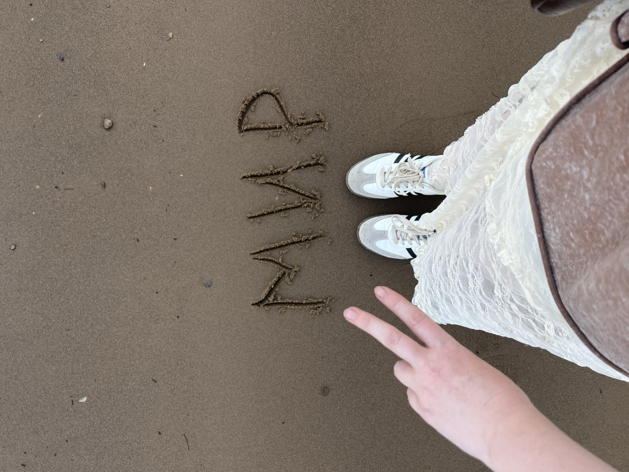 An overhead, medium shot captures a person's legs and feet standing on wet sand, with the word "МИР" (peace in Russian) written in the sand. The person is wearing white sneakers with black and blue stripes, and a long, white lace dress. Their left hand, with fingers extended in a peace sign, is visible in the lower left corner. The sand is dark brown and appears damp, with a few small pebbles and shells scattered across it. The background is entirely composed of this sandy surface. The scene suggests a location on a beach or riverbank, consistent with the Volga River embankment in Chaikovsky, Russia, where such writing in the sand and casual attire would be common.