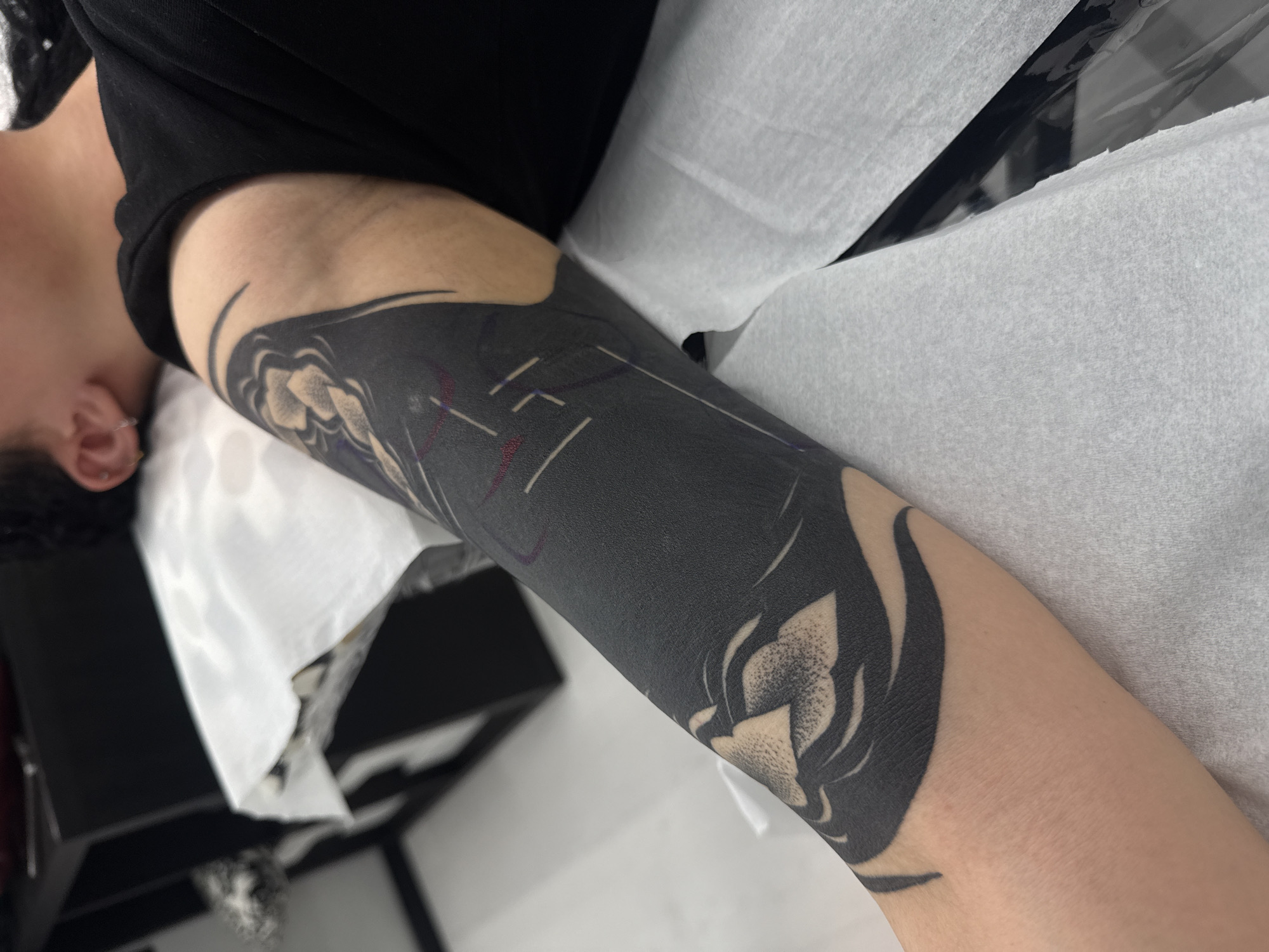 The image shows a close-up of a person's arm with a large, black tattoo covering a significant portion of the upper arm and possibly extending down. The tattoo features floral or petal-like designs incorporated into the black ink, creating a contrast between the skin and the tattoo. There appear to be faint guidelines or markings visible on the tattoo, possibly from the tattooing process. The arm is resting on what seems to be a white disposable surface, possibly in a tattoo studio or medical setting. The person is wearing a black shirt. The image does not have any features or details that would suggest it was taken in Sofia, Bulgaria. The image contains no information about the city of Sofia.