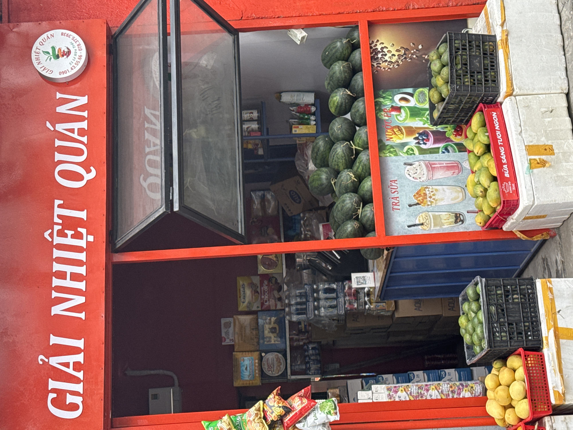 The image shows the exterior of a small convenience store in Đà Nẵng, Vietnam, identified by the red signage vertically displaying "GIẢI NHIỆT QUÁN" in white letters. A smaller, round sign above this contains the store's logo and phone numbers. The store appears to sell fresh fruits like watermelons and mangoes, which are stacked near the storefront. The interior shelves are stocked with various items, including drinks, snacks, and canned goods. The store features an open display case, and the overall setting appears to be a common scene in a Vietnamese urban environment.