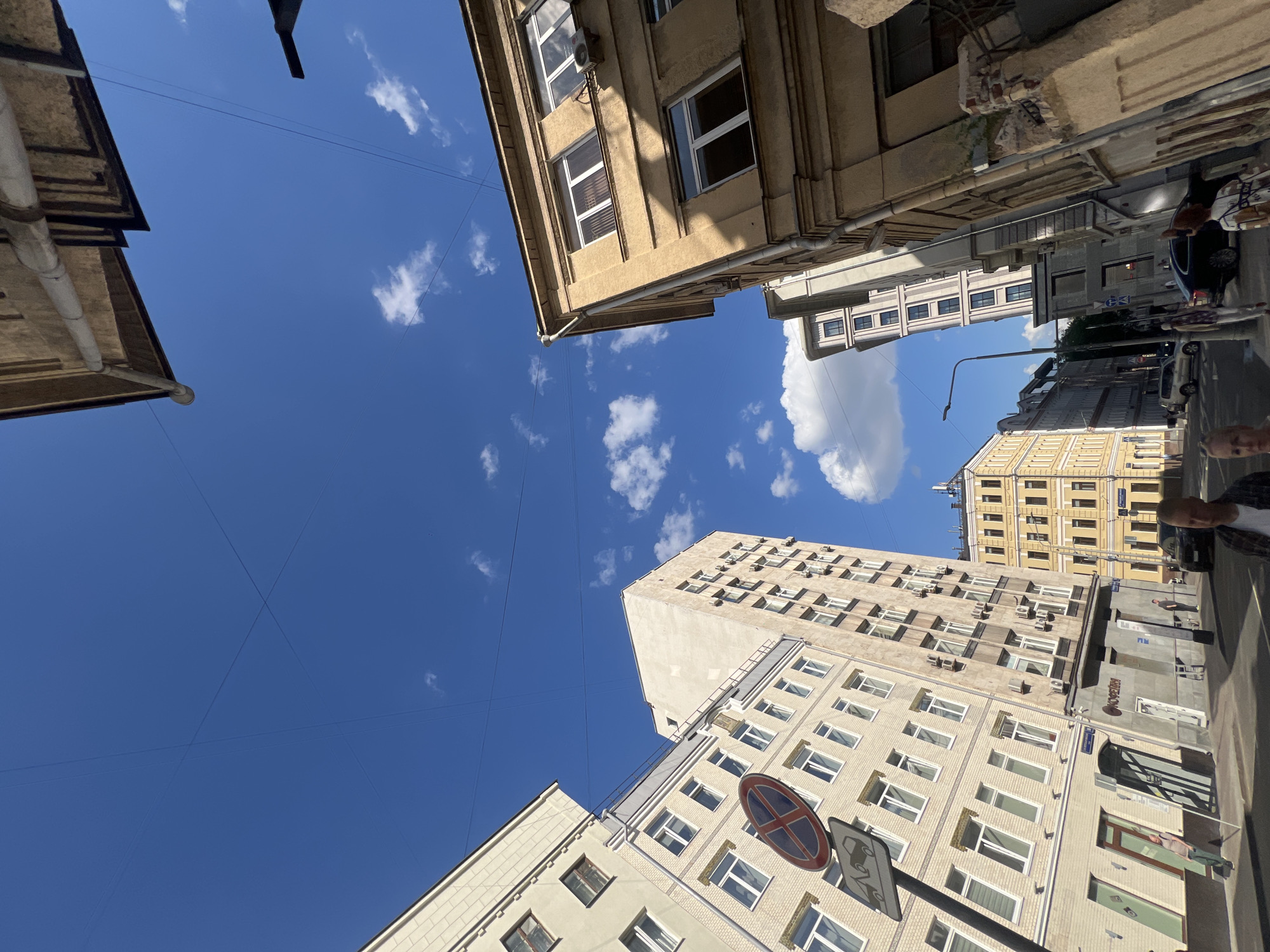 The image offers a worm's-eye perspective of a bustling city street in Moscow, Russia, looking upwards towards a vibrant blue sky. The sky dominates the center of the frame, featuring a few scattered white cumulus clouds and intricate patterns of thin overhead wires crisscrossing its expanse.
The scene is framed by various multi-story buildings, characteristic of urban architecture. These structures exhibit different shades of beige, cream, and light yellow, with numerous windows reflecting the bright daylight. Many buildings display visible air conditioning units mounted on their facades.
On the lower right, a street-level view reveals a typical urban environment with cars, including a dark sedan and a white vehicle, along with partially visible pedestrians. A person's head and shoulder are visible in the immediate foreground. Prominently featured on a pole is a red circular road sign with a diagonal white cross, commonly indicating "No Stopping" or "No Parking," and another sign below it depicting a car. On a building facade, the Cyrillic text "ОСЕРИЯ" is discernible. The sunlight suggests it is daytime with clear, pleasant weather.