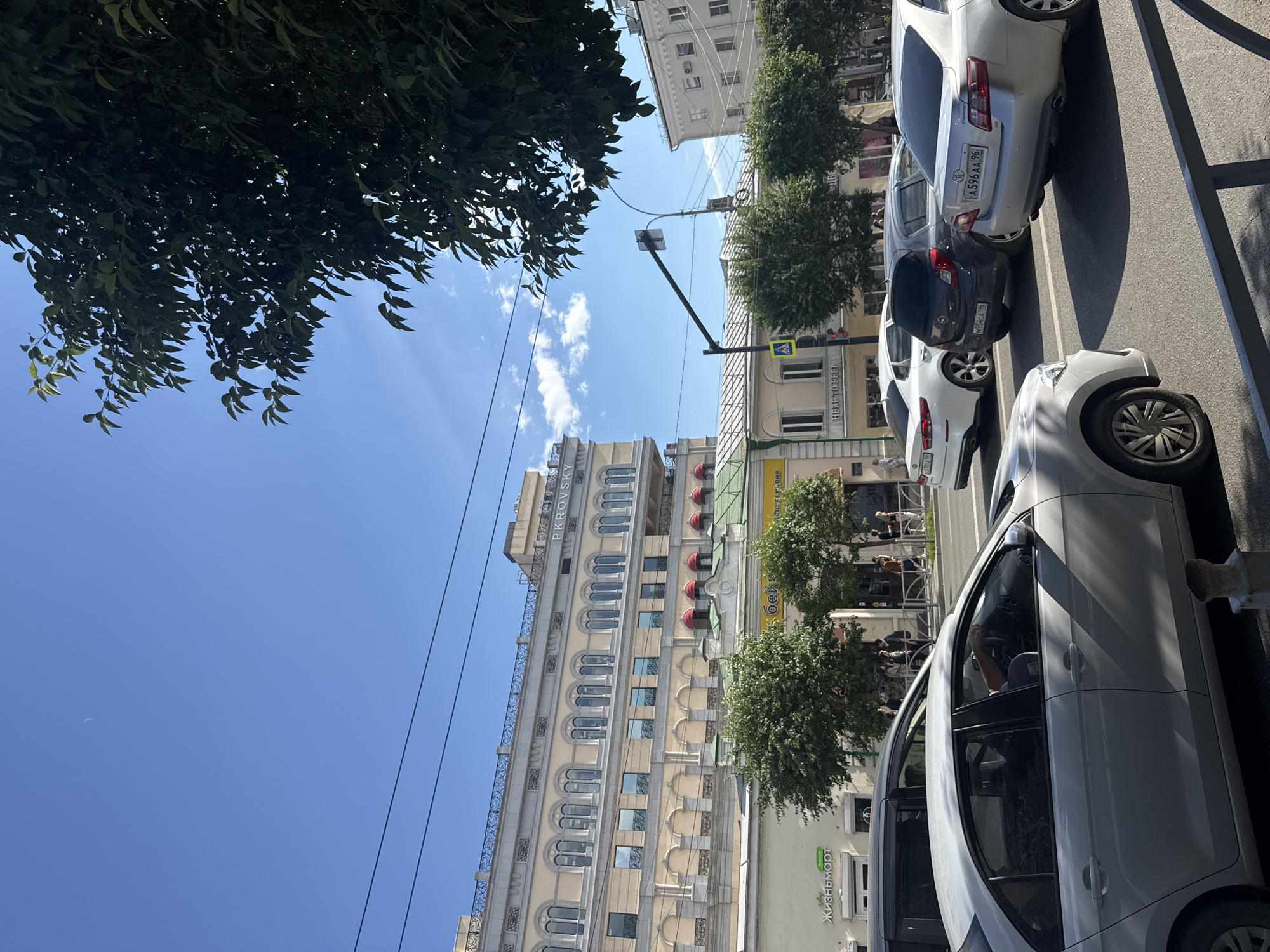 This media file depicts a street scene in Yekaterinburg, Russia, on a bright, sunny day. The perspective is from a low angle, looking up towards a clear blue sky with a few wispy clouds. Lush green trees with dense foliage overhang the scene from the left, casting shadows.
The main subjects are several cars parked along the side of a street, suggesting a typical urban environment. The cars are of various models and colors, including white, dark grey, and silver. One car, a silver sedan, has a license plate that is partially visible and appears to read "A 596 AA 96".
Behind the parked cars, several buildings are visible. One prominent building is a tall, light-colored structure with numerous arched windows and ornate architectural details. Text on this building reads "PKROVSKY". To the right of this building, there are other buildings with storefronts. One storefront has green signage with text that appears to be in Cyrillic script, possibly related to a business.
The time of day is clearly daytime, indicated by the bright sunlight and the clear sky. The weather appears to be warm and pleasant. There are no specific events captured, but the presence of parked cars and buildings suggests a normal day in the city. People are visible in the background, some walking on the sidewalk and others appearing to be inside or near the storefronts, though they are not the primary focus. The overall impression is one of a bustling urban street on a summer day.