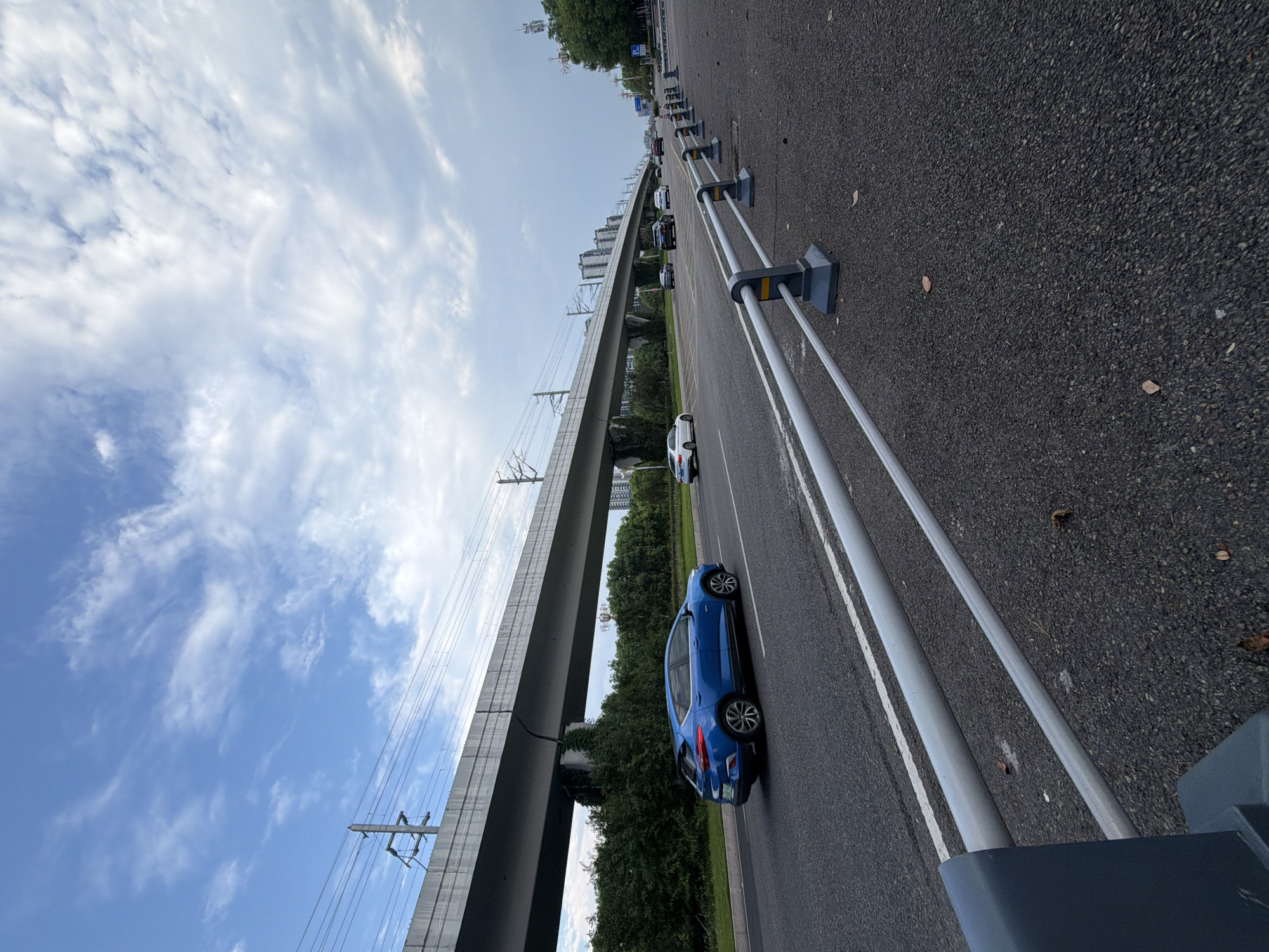 The image depicts a road with a train track overhead in Jiangjin, China. In the foreground, a grey metal guardrail runs along the side of the asphalt road. A blue hatchback car is traveling on the road, with several other cars visible further down the road. The road is flanked by trees and bushes, and in the background, there are buildings and a radio tower. The sky is cloudy with patches of blue. The overall scene suggests an urban or suburban environment with active transportation infrastructure.