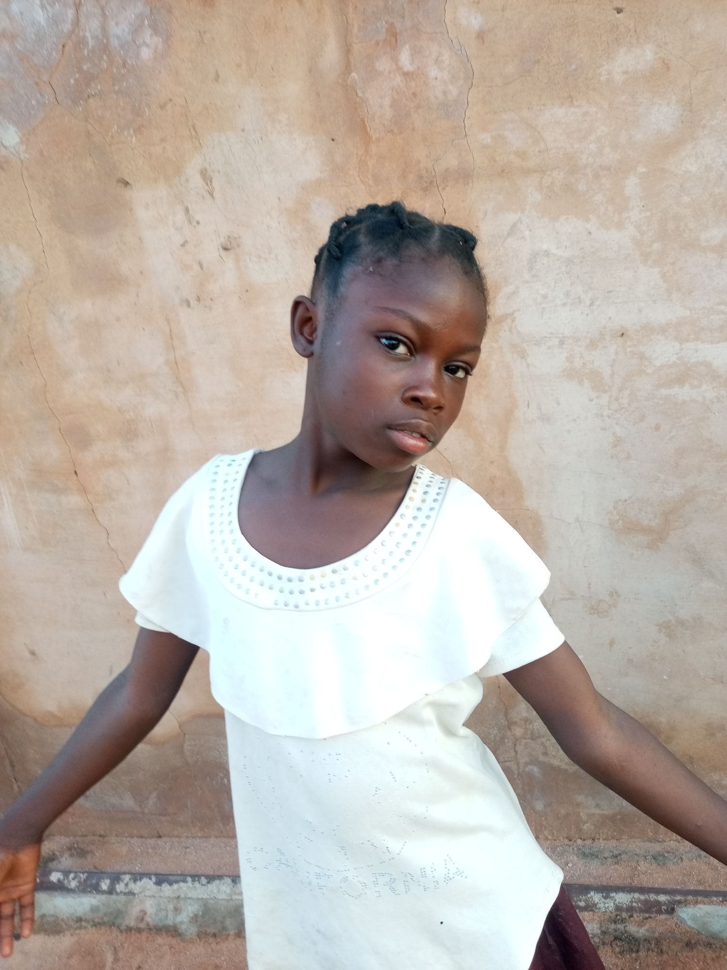 A medium shot photograph captures a young Black girl, likely under ten years old, standing against a textured, light brown wall. The girl has dark skin, black braided hair styled away from her face, and dark eyes. She is wearing a light-colored dress with a ruffled neckline adorned with small, iridescent beads or sequins. The dress appears to be made of a textured fabric and has short, puffed sleeves. Her arms are extended slightly to the sides. The background wall shows signs of age and weathering with visible cracks and patches of lighter discoloration. A horizontal element, possibly a ledge or the top of a fence, runs across the lower part of the image. The lighting suggests an outdoor setting with natural light illuminating the scene from the front.