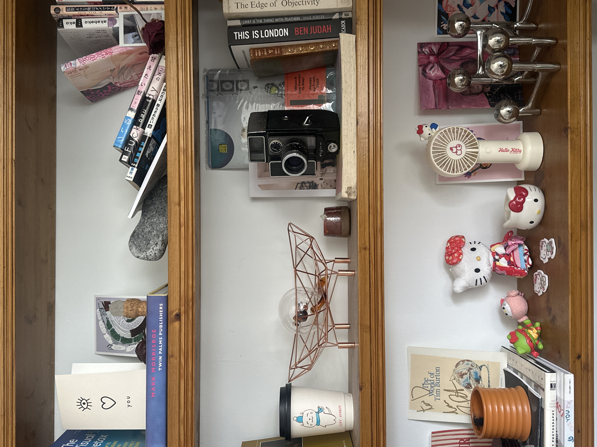 The image depicts three adjacent shelves filled with assorted items. The shelves, constructed of wood with a light brown finish, are arranged vertically.
The leftmost shelf contains a collection of books, some with Japanese text, and a grey rock. Also visible are a card with an eye and heart symbol and a book titled "MARK MORRISROE TWIN PALMS PUBLISHERS".
The middle shelf features more books, including "The Edge of Objectivity," "THIS IS LONDON BEN JUDAH," and "GRIEF IS THE THING WITH FEATHERS MAX PORTER". A black Fujica camera sits atop a pile of photos or postcards, and a copper-colored wire sculpture and a coffee cup are positioned below.
The rightmost shelf holds various objects, including a silver decorative item resembling branching arms, a painting with a pink bow, a Hello Kitty fan, a Hello Kitty head and doll, several small figurines, a signed copy of "The World of Tim Burton", and an orange plant pot.
The setting appears to be indoors, perhaps within a private residence in the Folkestone and Hythe District, United Kingdom, given the presence of books and personal items.