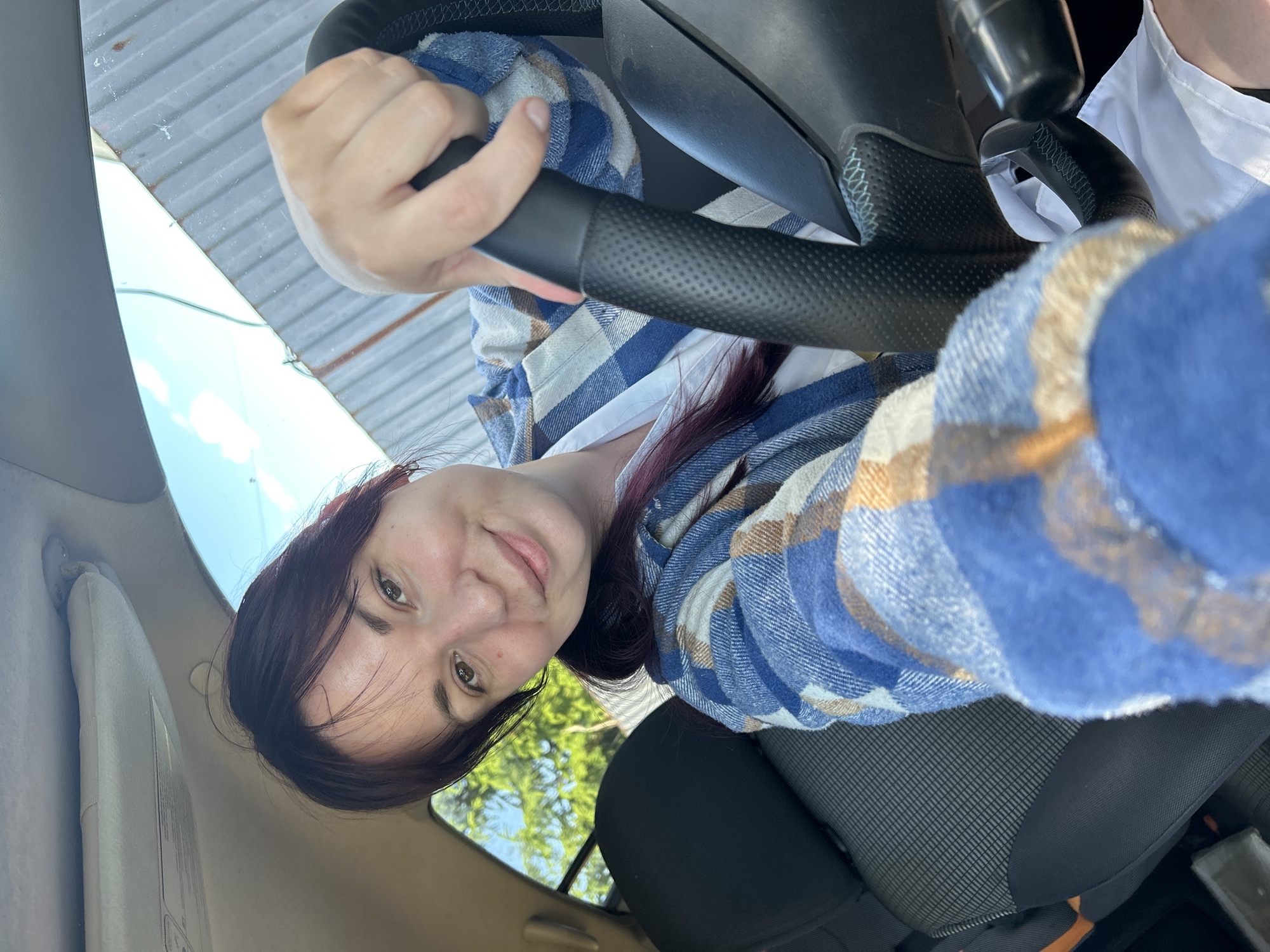 A young woman with dark hair, wearing a blue and tan plaid shirt over a white top, is seated in the driver's seat of a car. She is holding the steering wheel with her right hand and looking at the camera with a slight smile. The interior of the car is visible, including the headrest of the passenger seat and the steering wheel. Outside the car, a corrugated metal fence and trees are visible under a blue sky with scattered clouds. The location is identified as Фоки, Russia.