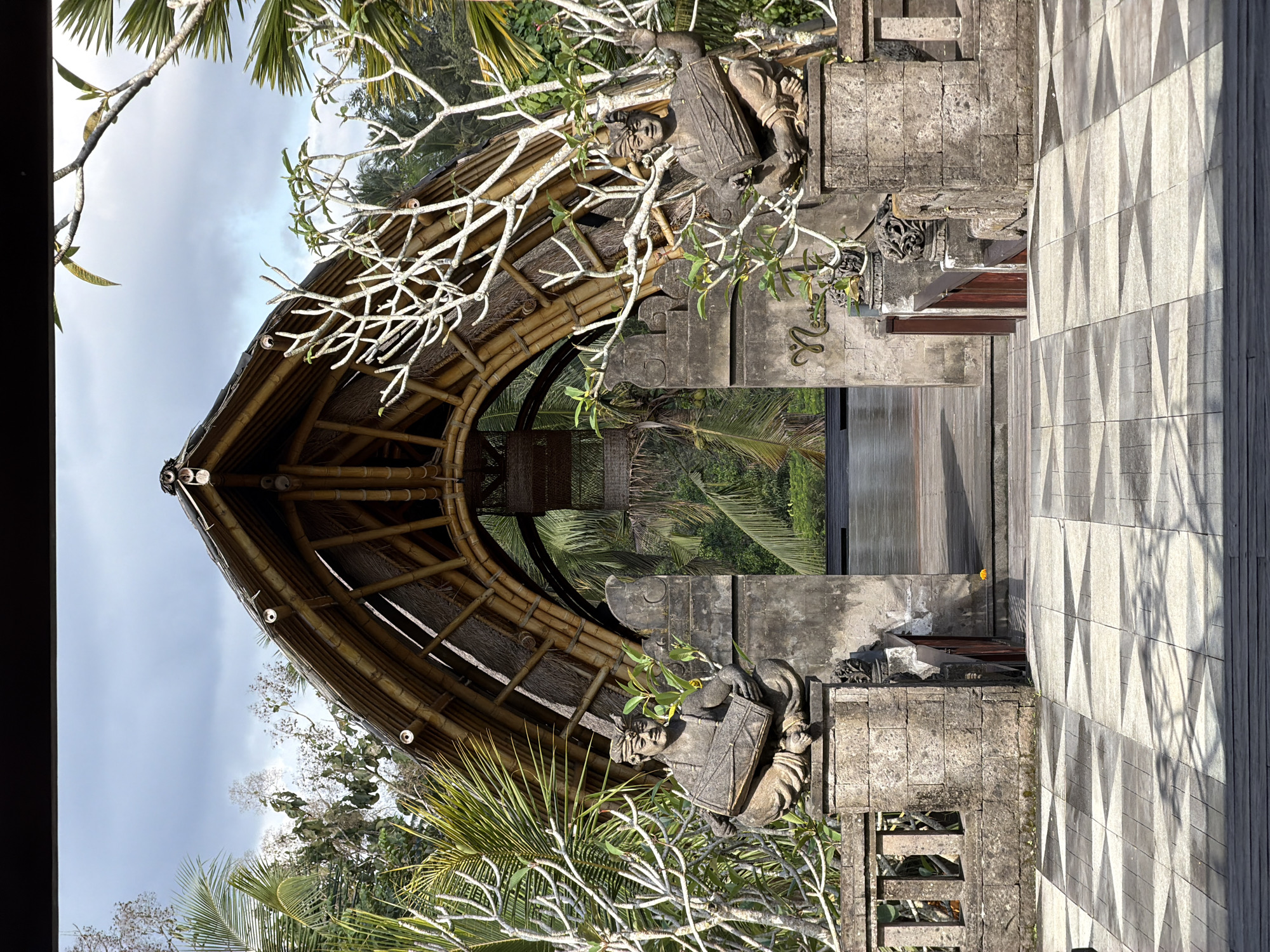 The image depicts an ornate entranceway to a resort or cultural site in Tegallalang, Indonesia. The entrance is framed by a large bamboo archway covered with thatch, curving upwards to a peak. On either side of the stone pathway leading through the archway stand two weathered stone statues of figures seated cross-legged and playing a cylindrical drum. These statues are positioned on stone pedestals that are part of a low stone wall bordering the pathway.
The pathway itself is paved with a geometric pattern of square and triangular tiles in shades of light and dark grey. Behind the statues and the archway, lush green tropical foliage, including palm trees, is visible. The sky above is partly cloudy, with patches of blue visible. The overall impression is one of an exotic and culturally rich environment, typical of Bali. The entrance appears to lead to an open area, possibly a pool or a gathering space, with a wooden deck visible beyond the archway. The lighting suggests it is daytime, with shadows cast on the tiled pathway.