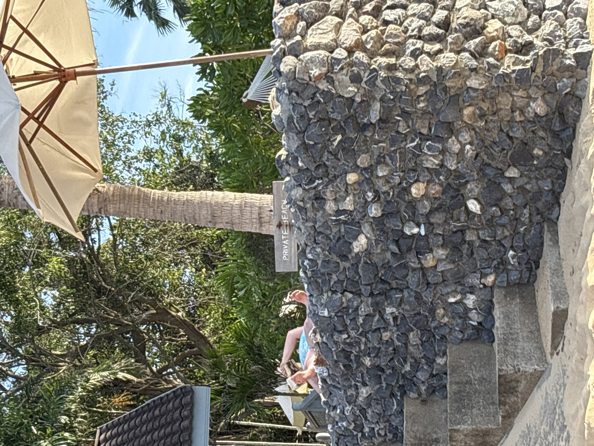 Baan Bo Phut, Thailand: A medium shot depicts a scene featuring a stone wall covered in various shades of grey rocks, adjacent to a set of weathered stairs, also grey. Attached to the wall is a sign that reads "PRIVATE BEACH." The stairs lead towards a seated individual, partially obscured, whose bare legs are visible, wearing a light blue shirt. To the left of the wall and stairs, trees and other foliage are seen, creating a background of green. A white parasol is also visible in the upper left of the image.