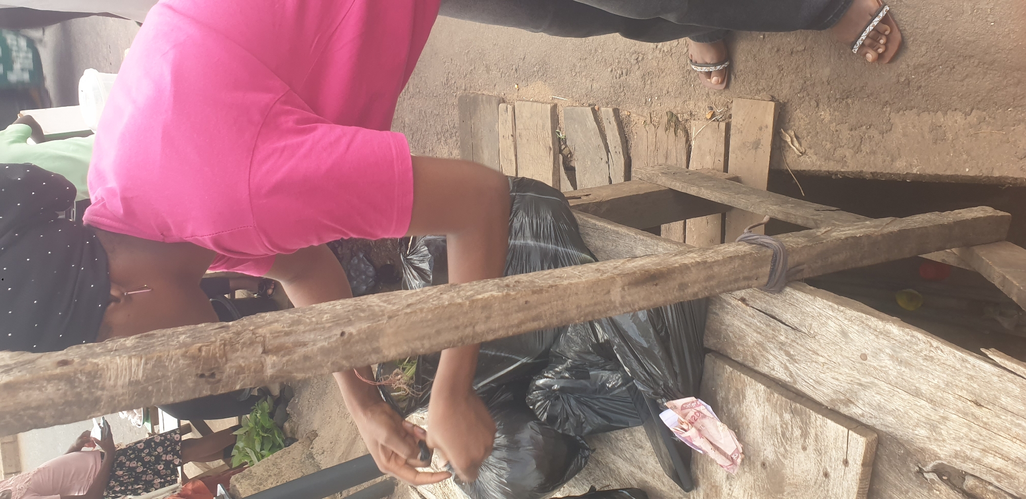 This media file captures a scene from Barakin Lerawi, Nigeria, possibly at an outdoor market. The primary subject is a person wearing a bright pink shirt and a black headscarf with white polka dots, leaning over a wooden structure. They are actively engaged in placing black plastic bags filled with items into the structure. Another person, partially visible in the background, wears a light green shirt. To the right, a person's feet are visible, wearing decorative sandals. The setting appears to be rustic, with rough wooden planks forming a sort of bin or stall. One of the black bags contains crumpled pieces of what appears to be currency, with the numbers "20" and "20" visible on one fragment. The lighting suggests it is daytime, and the ground appears to be dirt. The overall atmosphere seems to be one of commerce or perhaps a community distribution point.