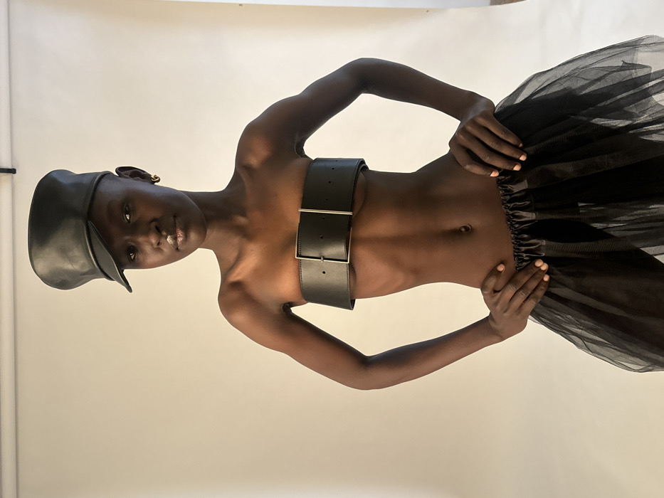 A dark-skinned individual, identified as a model, stands centrally in an indoor photography studio located in Paris, France. The subject wears a black leather military-style cap, a wide black leather belt fashioned as a strapless top with a prominent silver buckle, and a black sheer tulle skirt. A small gold hoop earring is visible in the subject's right ear. The individual has a shaved head and maintains direct eye contact with the viewer, exhibiting a neutral expression. Both hands are positioned on the hips. The background is a plain, light-colored seamless backdrop, typical of a professional studio setup, with consistent, even lighting illuminating the scene.