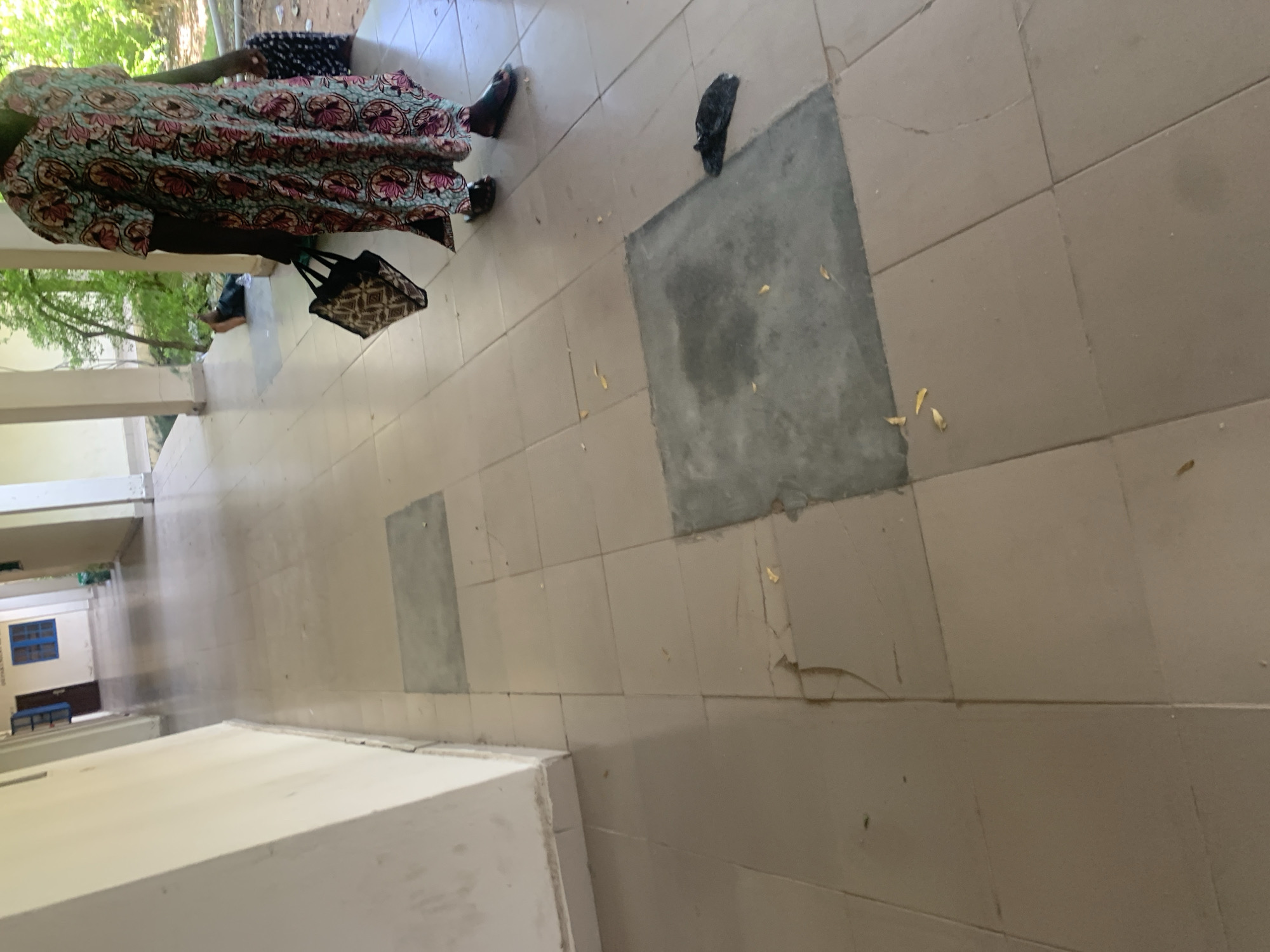 The media file shows a close-up view of tiled flooring in what appears to be an outdoor or semi-outdoor walkway or corridor. A person, seen from the knees down, is wearing a patterned dress and sandals. They are carrying a patterned bag in their right hand. In the background, there are trees and vegetation visible. The floor tiles are a light beige color, with some darker, square patches that may be repairs or distinct sections of flooring. There is also a black object lying on the floor near one of the darker patches. Further down the corridor, a blue-framed window and a dark blue bench can be seen. The lighting suggests it is daytime. The setting appears to be in Jalingo, Nigeria.